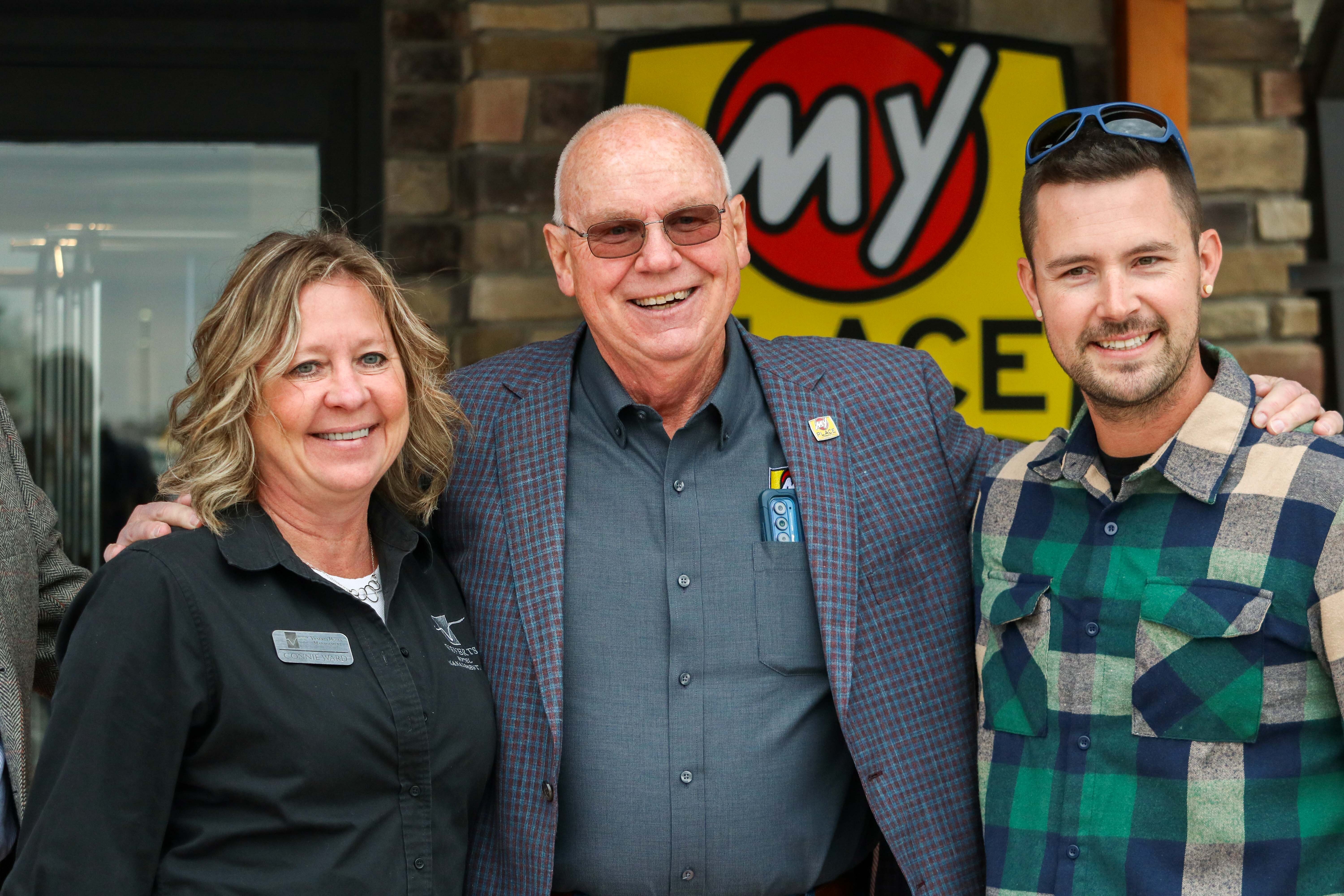 A blond woman in a black shirt, an older man with a gray jacket and glasses, and a younger man in a green flannel button-up pose for the camera outside the My Place Hotel in Mitchell, SD.