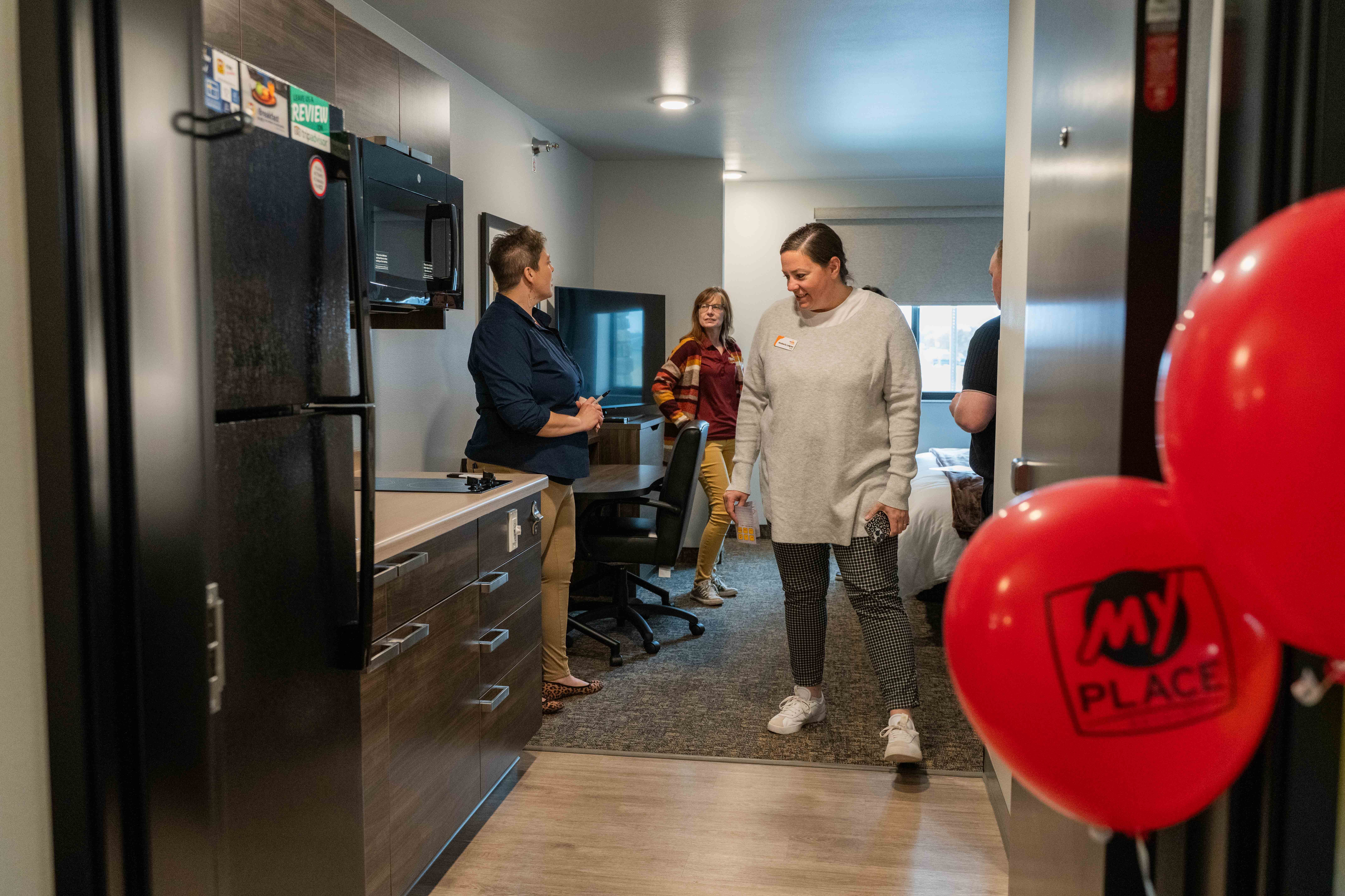 A woman in a dark blue shirt and khakis talks to three people inside a My Place Hotel room.