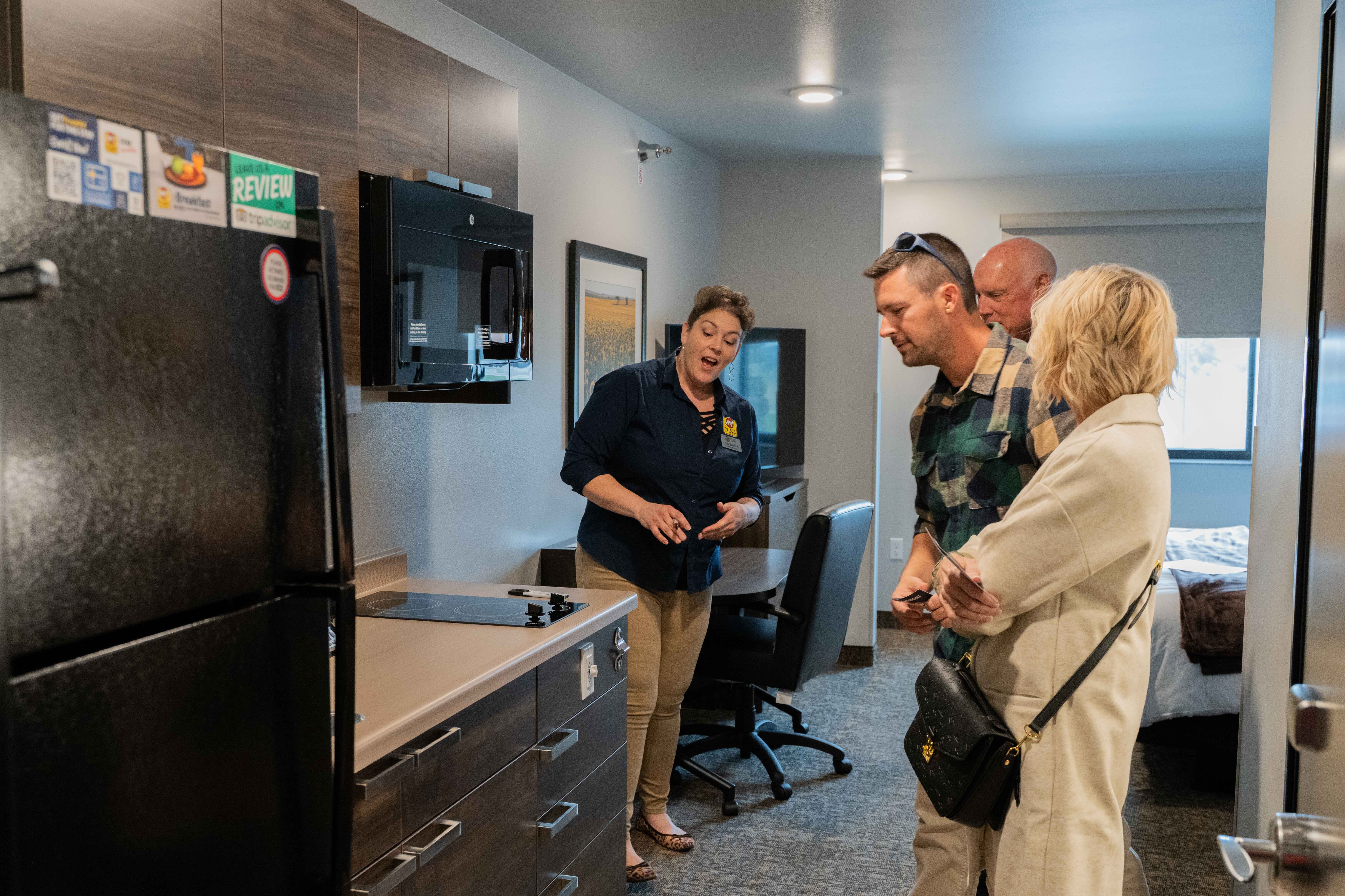 A woman wearing a dark blue button-down shirt and khakis stands in a My Place Hotel room with three people as she shows them a feature on the room's cooktop.