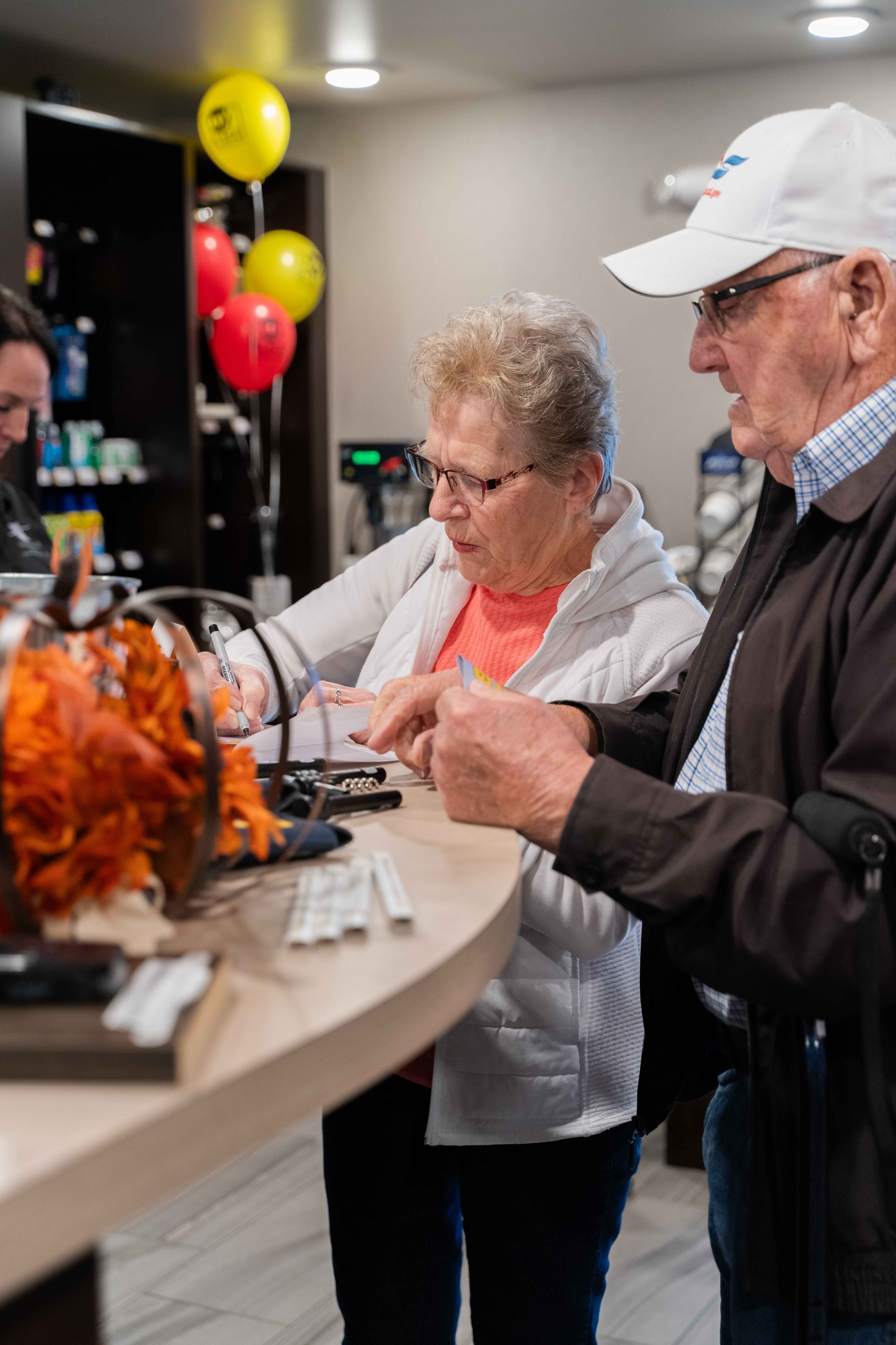 A woman wearing a white jacket and a man in a white hat write on paper while standing at the front desk inside My Place Hotel in Mitchell, SD.