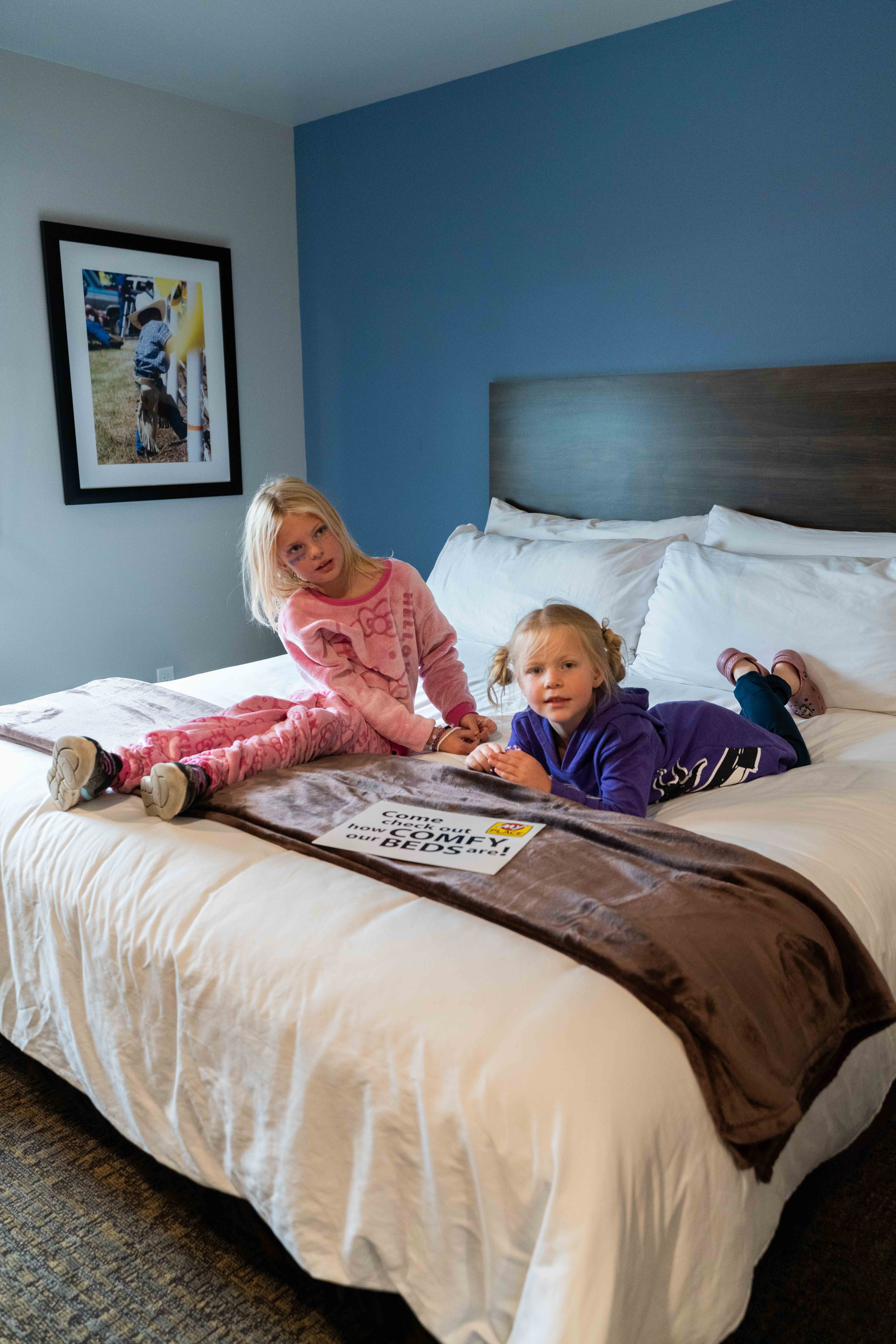 Two young girls wearing pink and purple pajamas sit on a My Place Hotel bed with a blue wall in the background.