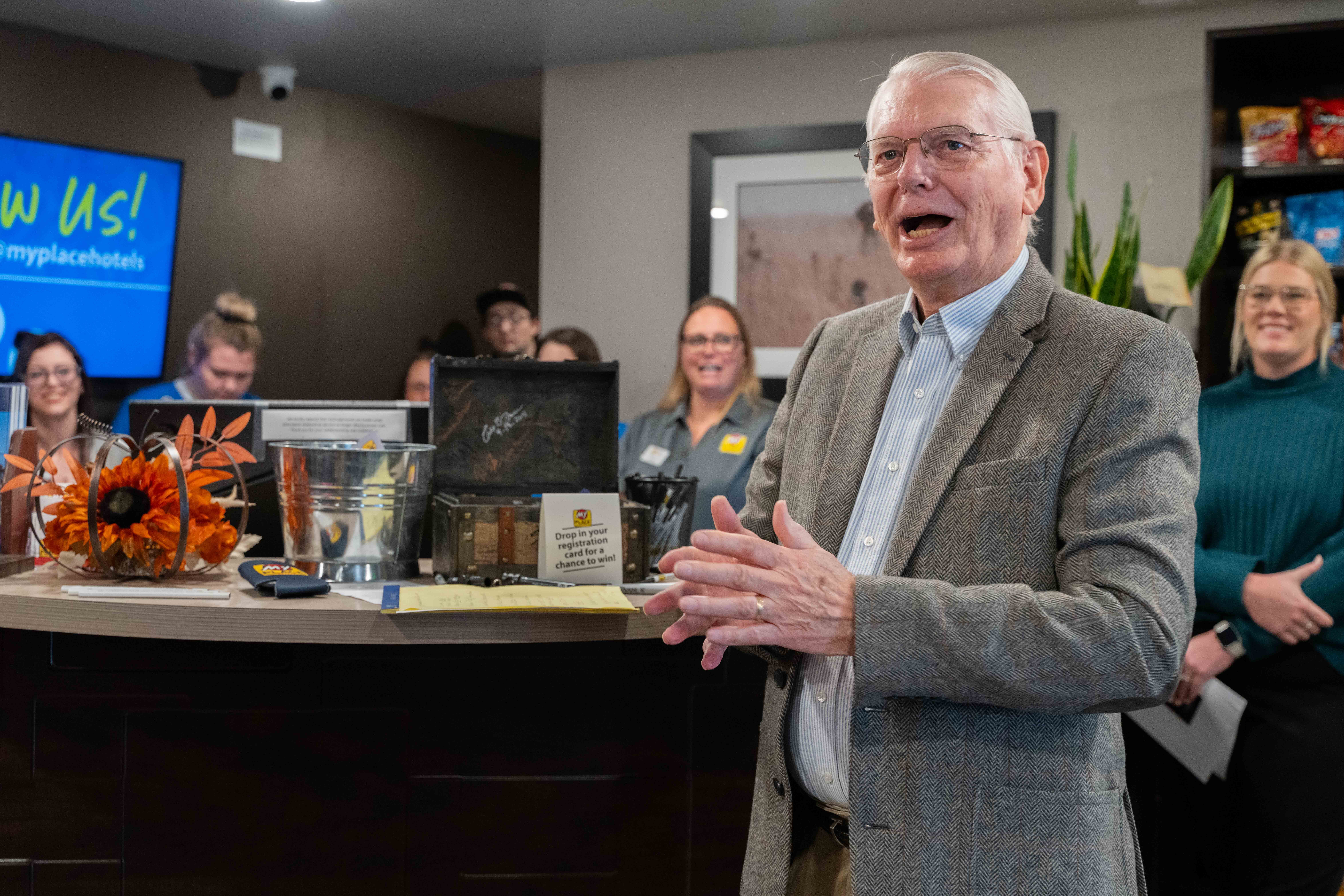 A man with a gray suit jacket and glasses addresses an off-camera crowd while seven people look on behind him inside a hotel lobby.
