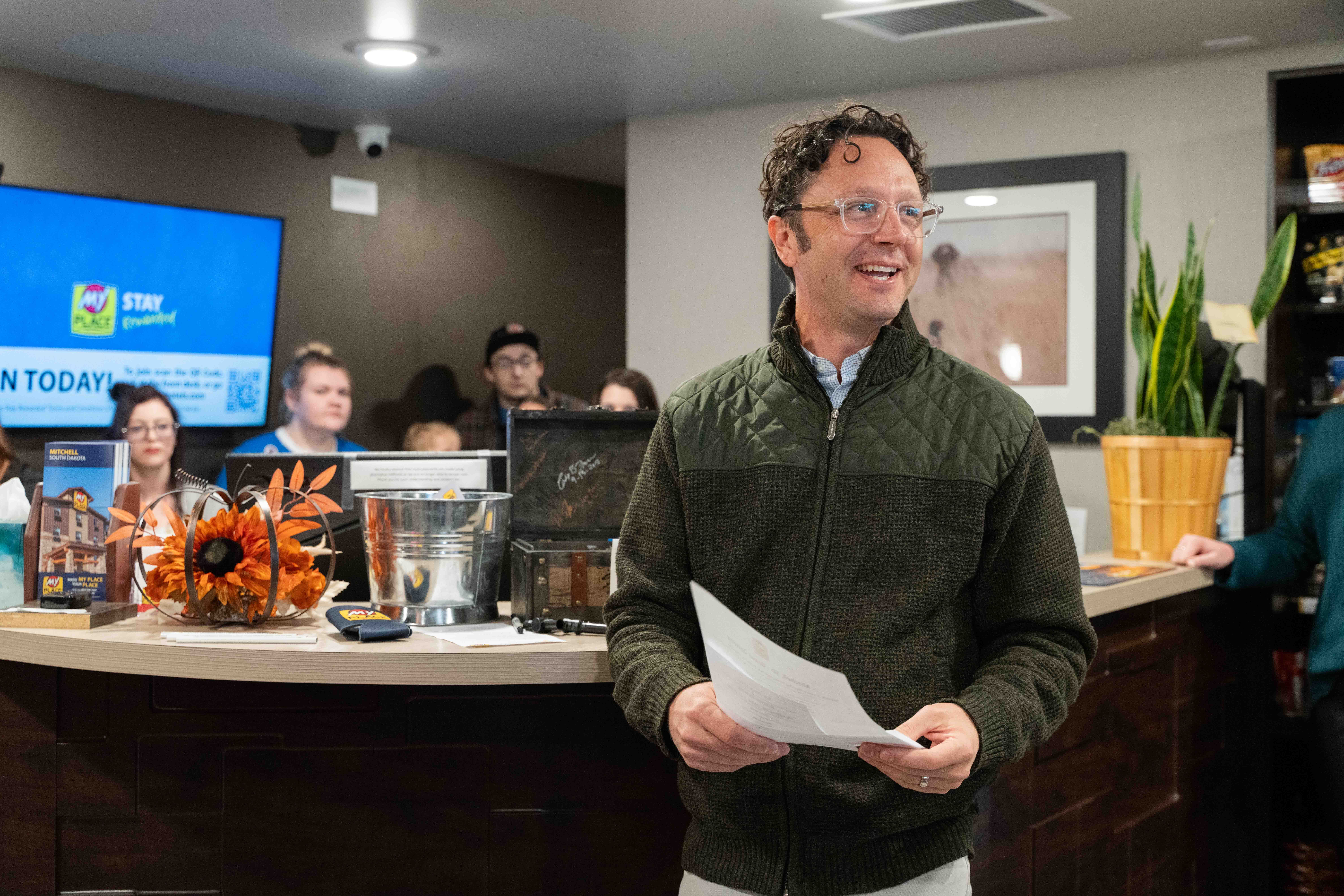 A man in a green zip-up sweater and glasses smiles at an off-camera crowd while five people look on behind him inside a hotel lobby.