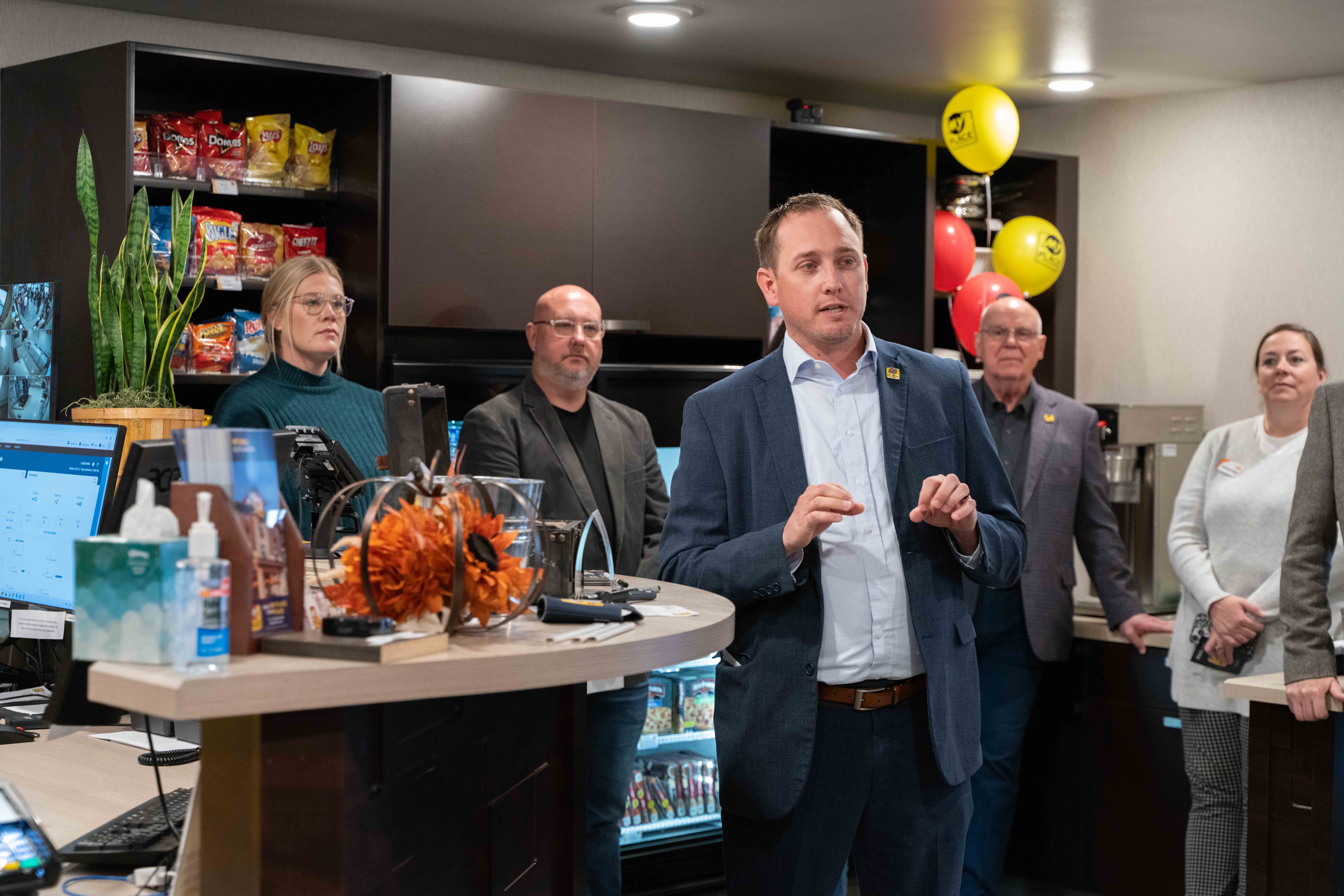 A man in a light blue shirt and dark blue suit jacket addresses an off-camera crowd while four people look on in the background inside a hotel lobby.