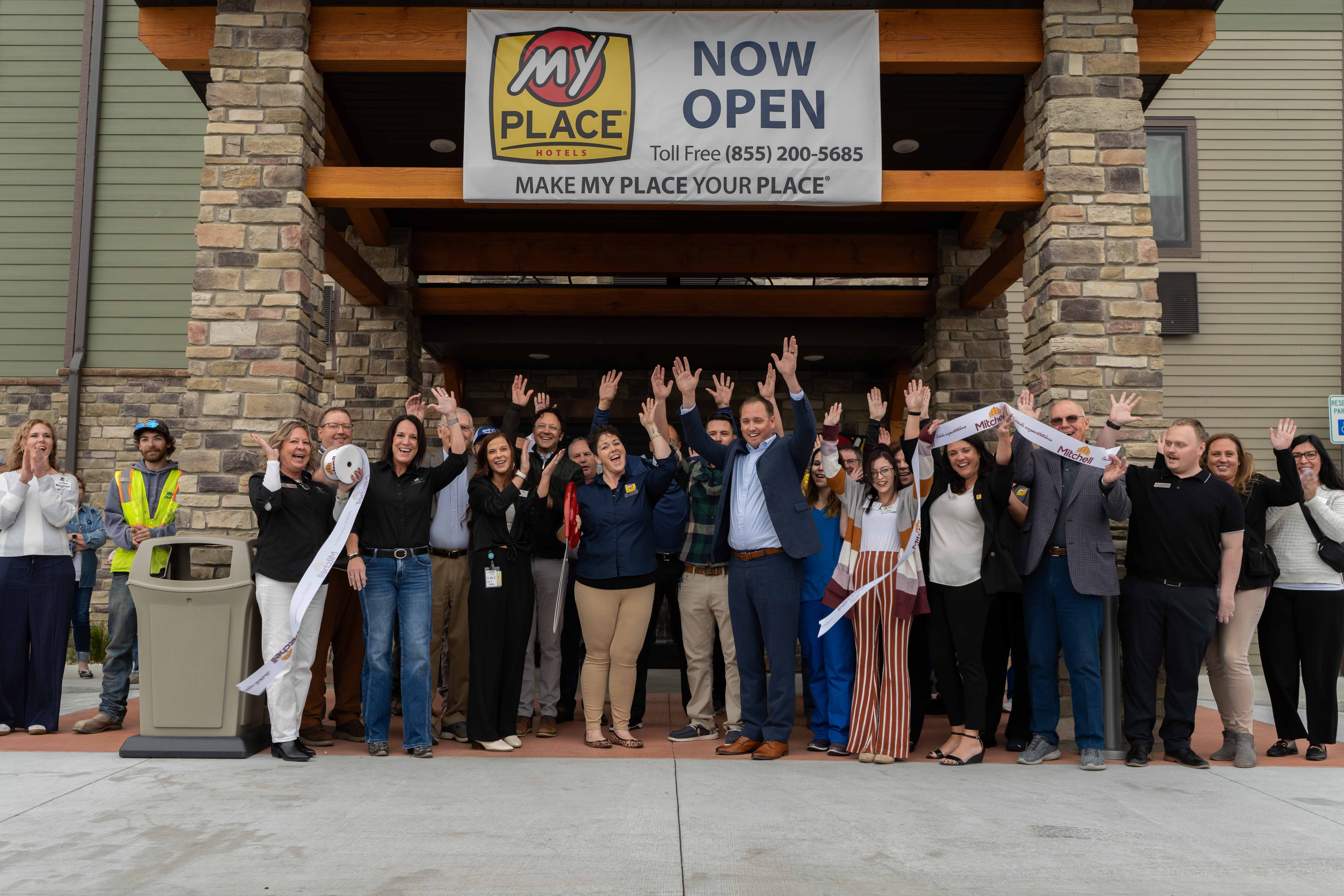 A large group of people standing outside under a Now Open sign throw their hands in the air in celebration following a ribbon cutting.