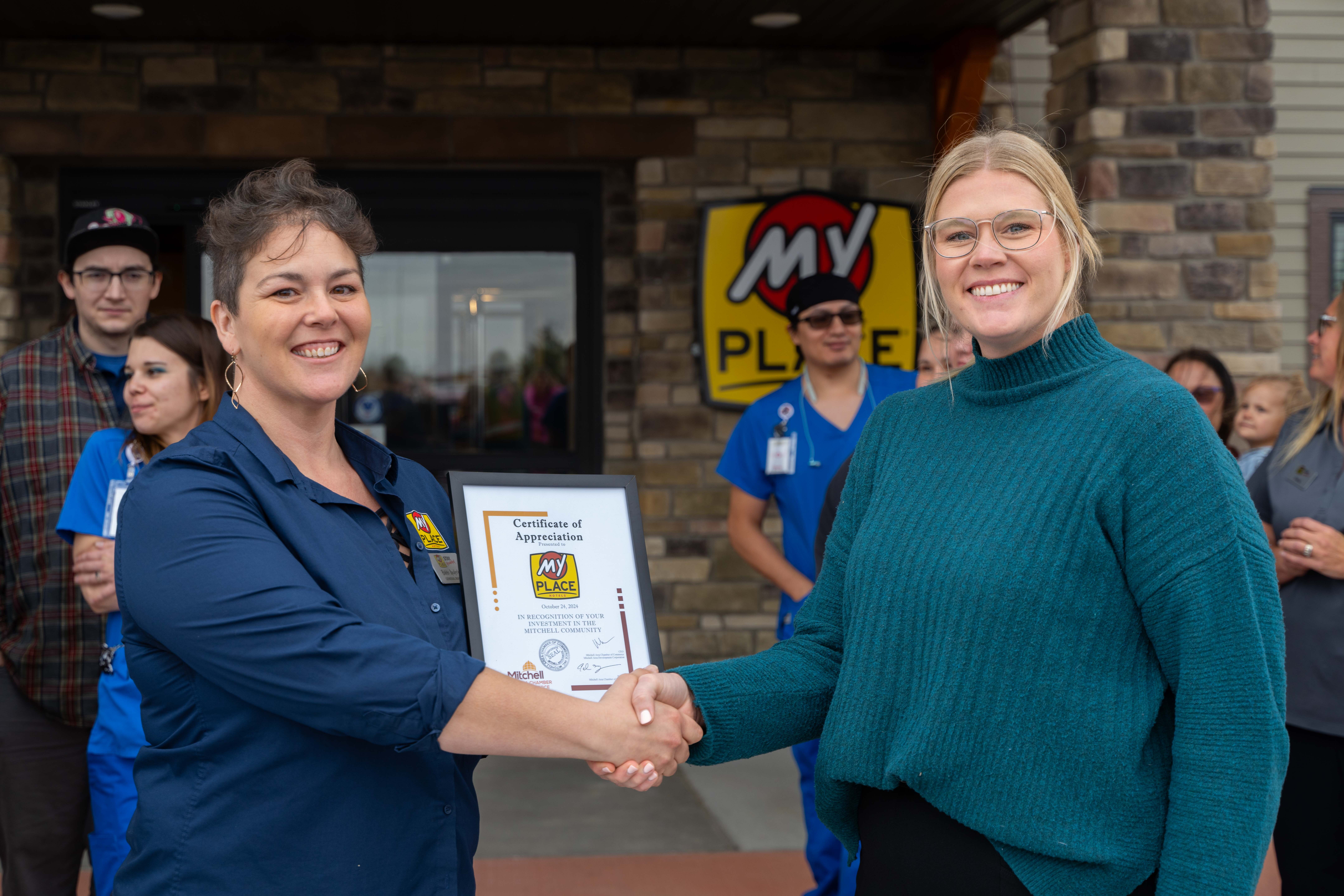 A woman with short, dark brown hair and a navy blue shirt shakes hands with a blonde woman wearing glasses and a teal sweater, both smiling at the camera.