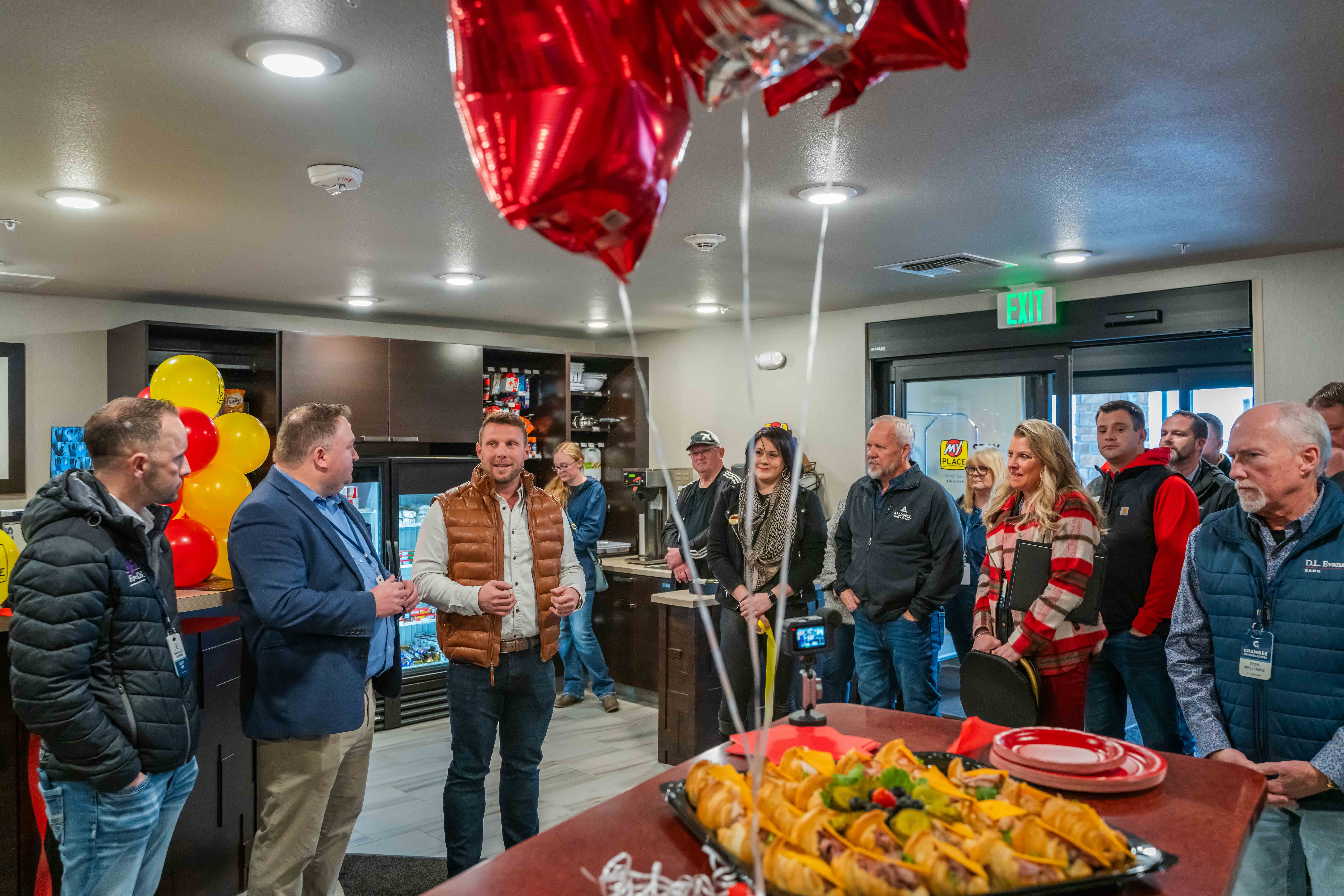 A group of people inside a My Place Hotel listening to Ryan Rivett speak during a grand opening celebration
