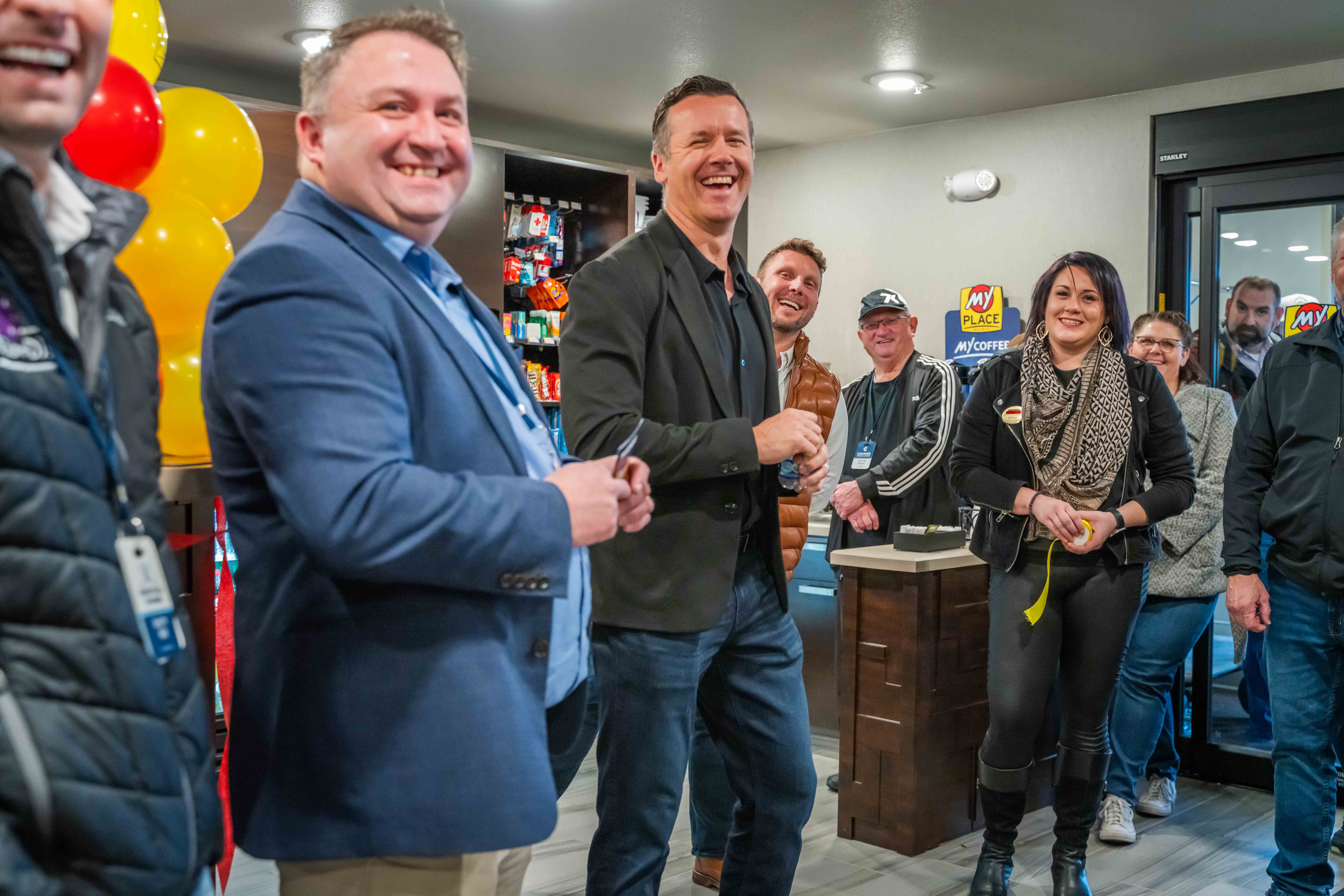 a group of people laughing at a joke inside a My Place Hotel during a grand opening celebration