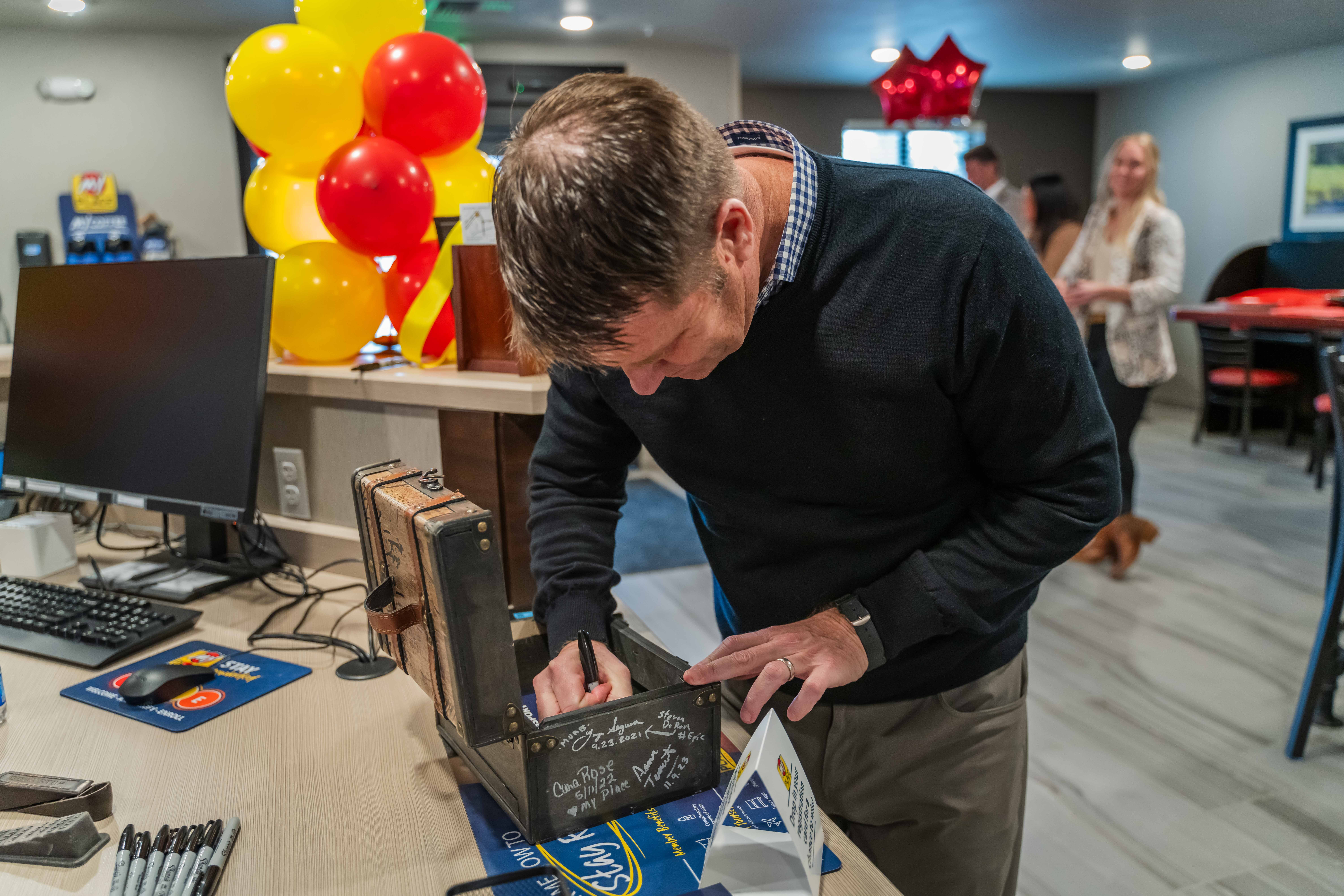 A man signing a decorative suitcase during a My Place Hotels grand opening celebration