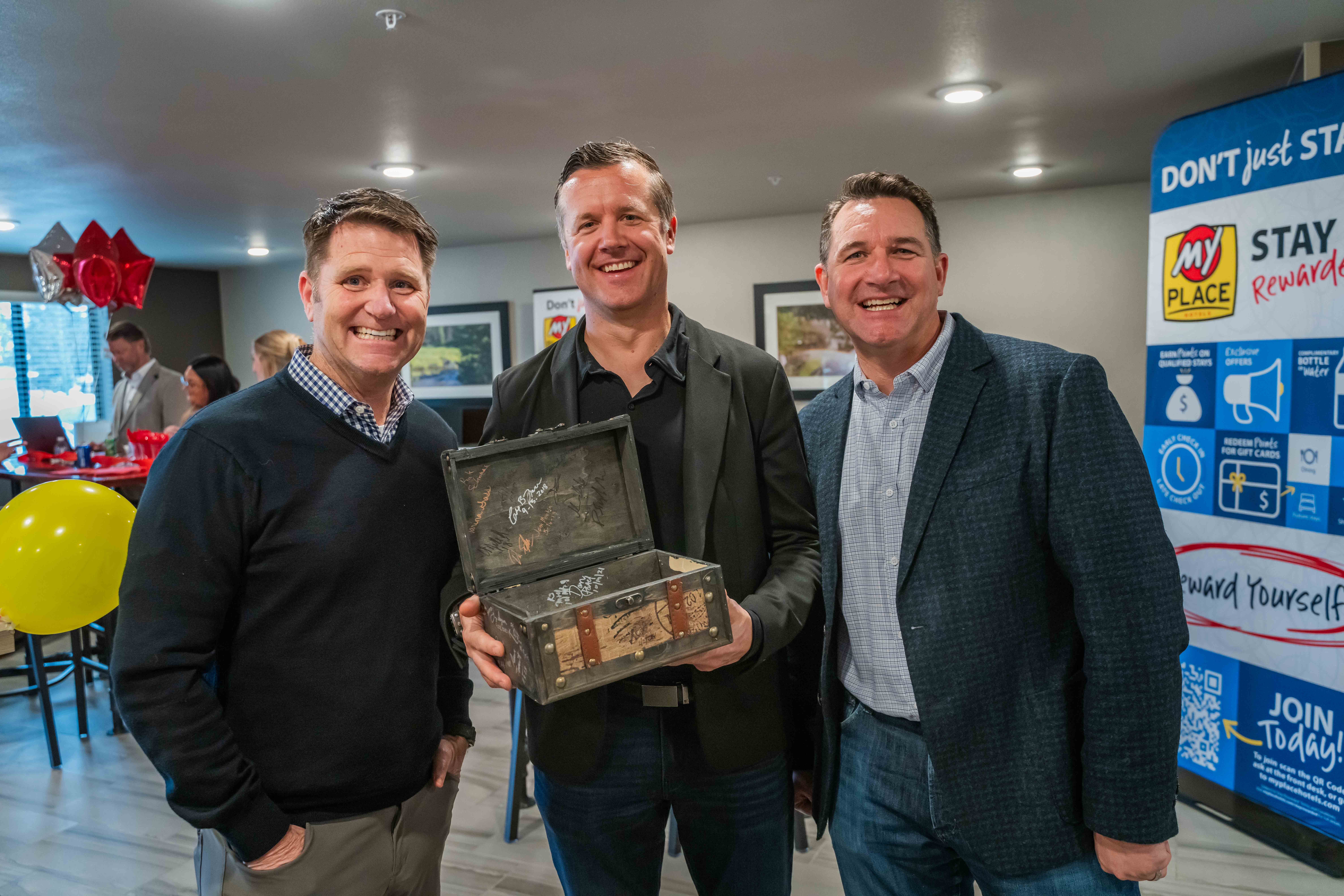 Three men standing with a decorative suitcase during a grand opening celebration in a hotel lobby