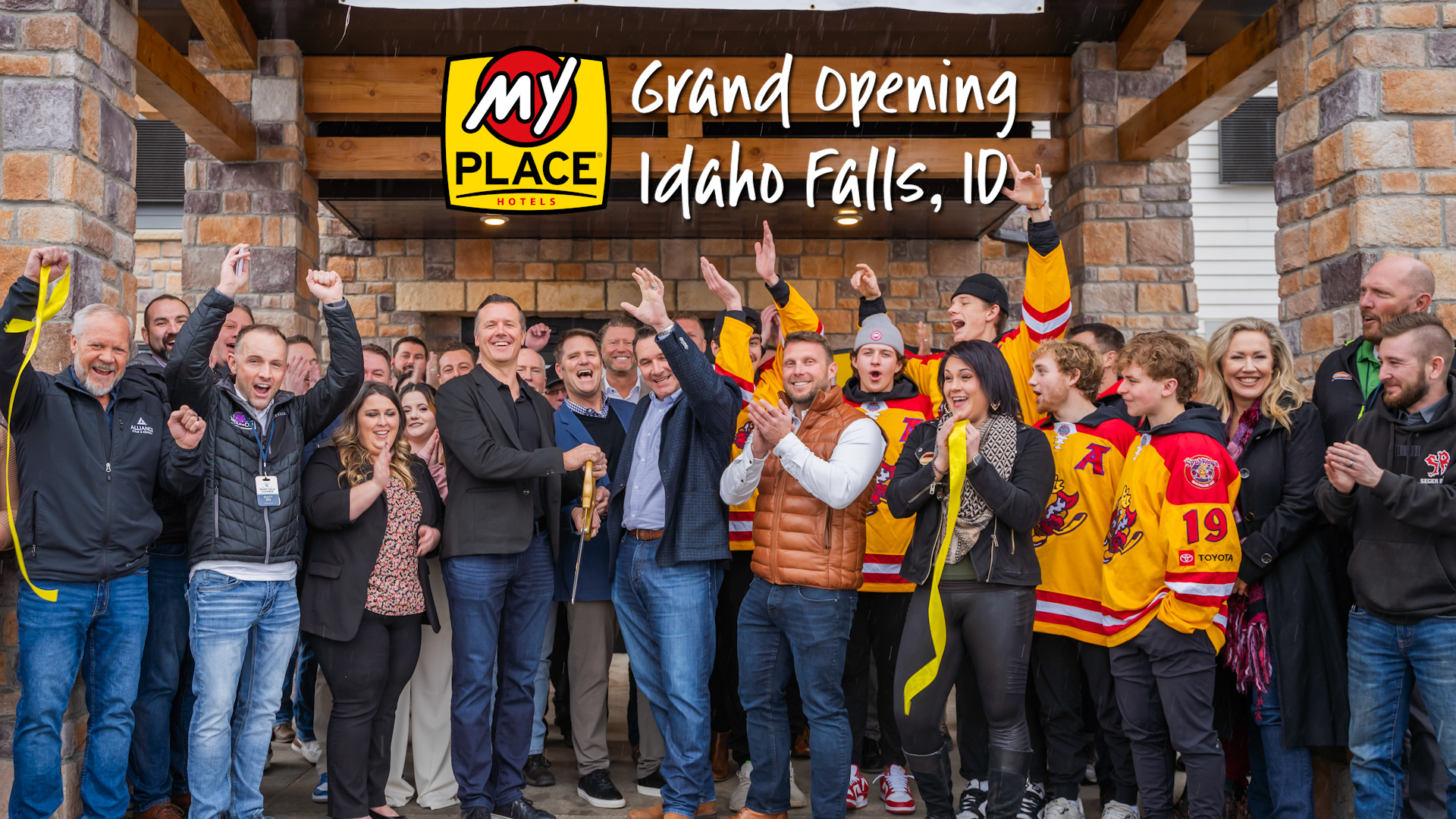 Group of people celebrating a My Place Hotels ribbon cutting with text at the top reading Grand Opening Idaho Falls, ID