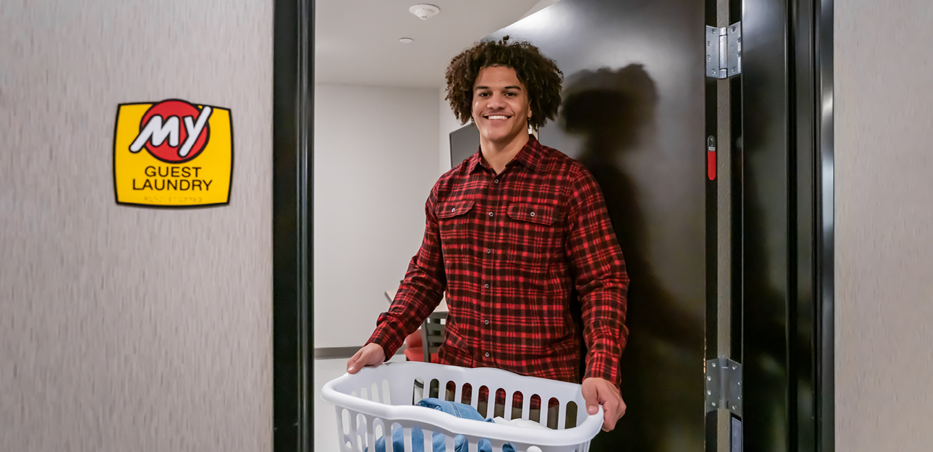 A man smiles as he stands in the doorway to the laundry facility holding a laundry basket.