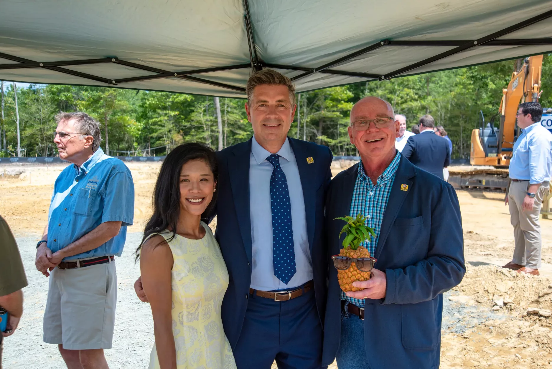 Three people smile and pose for the camera under a tent with construction equipment in the background.