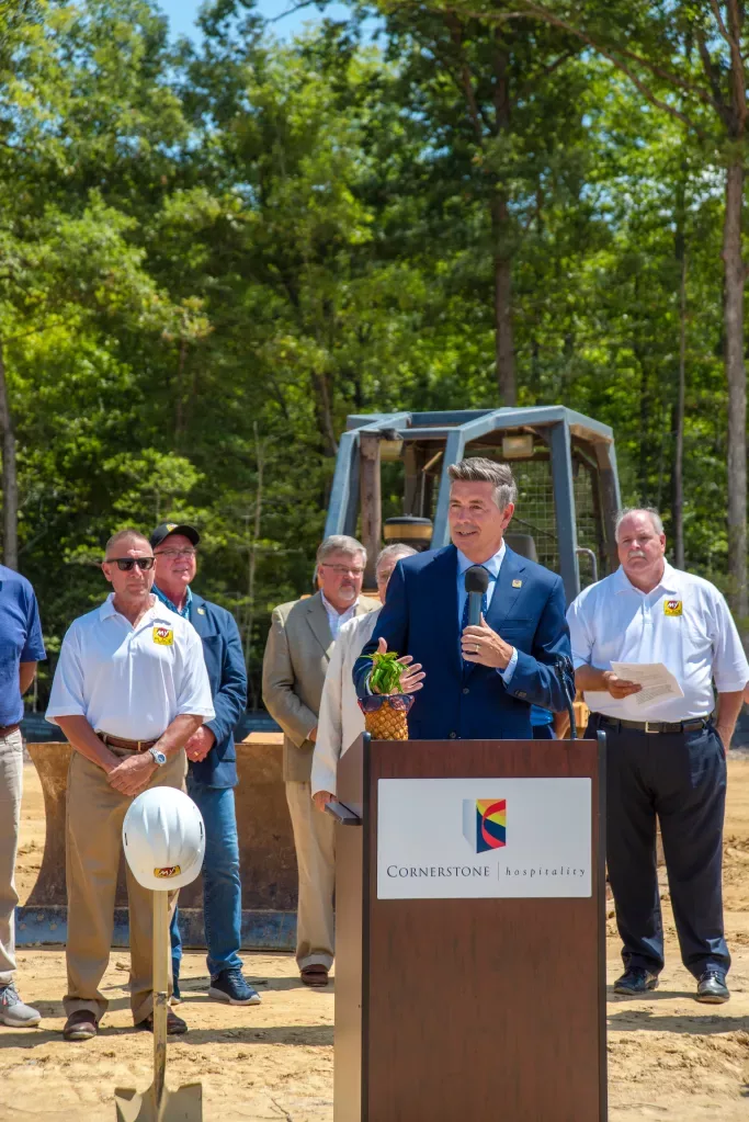 A man holds a microphone and addresses an off-camera group of people outdoors with several men in the background.