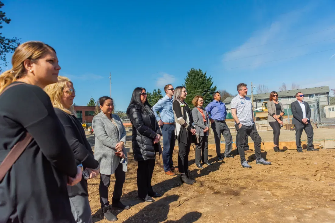 About a dozen people watch an off-camera speaker surrounded by fresh dirt at the groundbreaking ceremony in Vancouver, Washington.