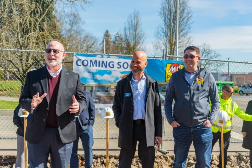A man addresses an off-camera crowd while two other men look on during the groundbreaking ceremony in Vancouver, Washington.