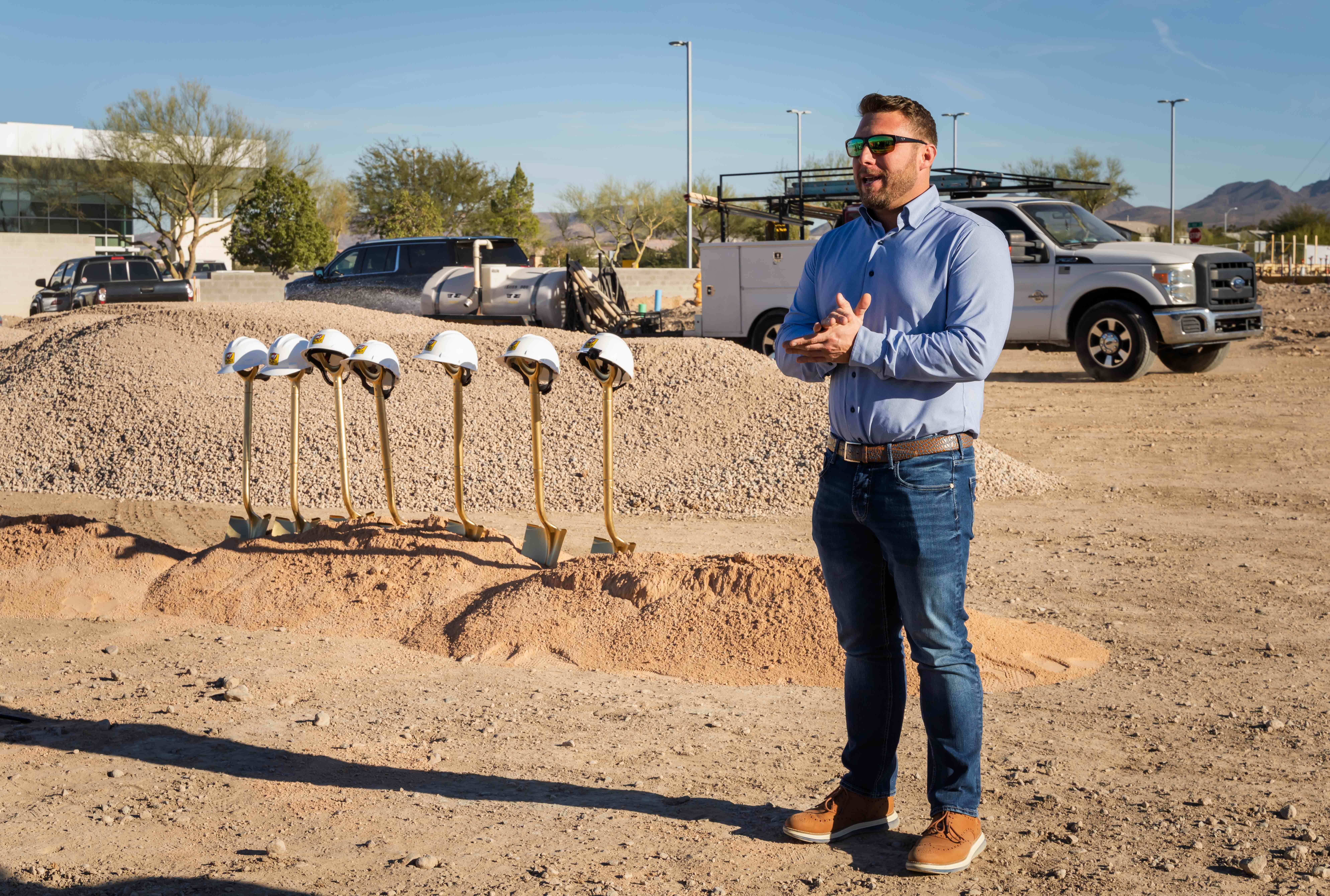 My Place CEO Ryan Rivett addressing crowd at groundbreaking construction site with gold shovels in and dirt in the background.