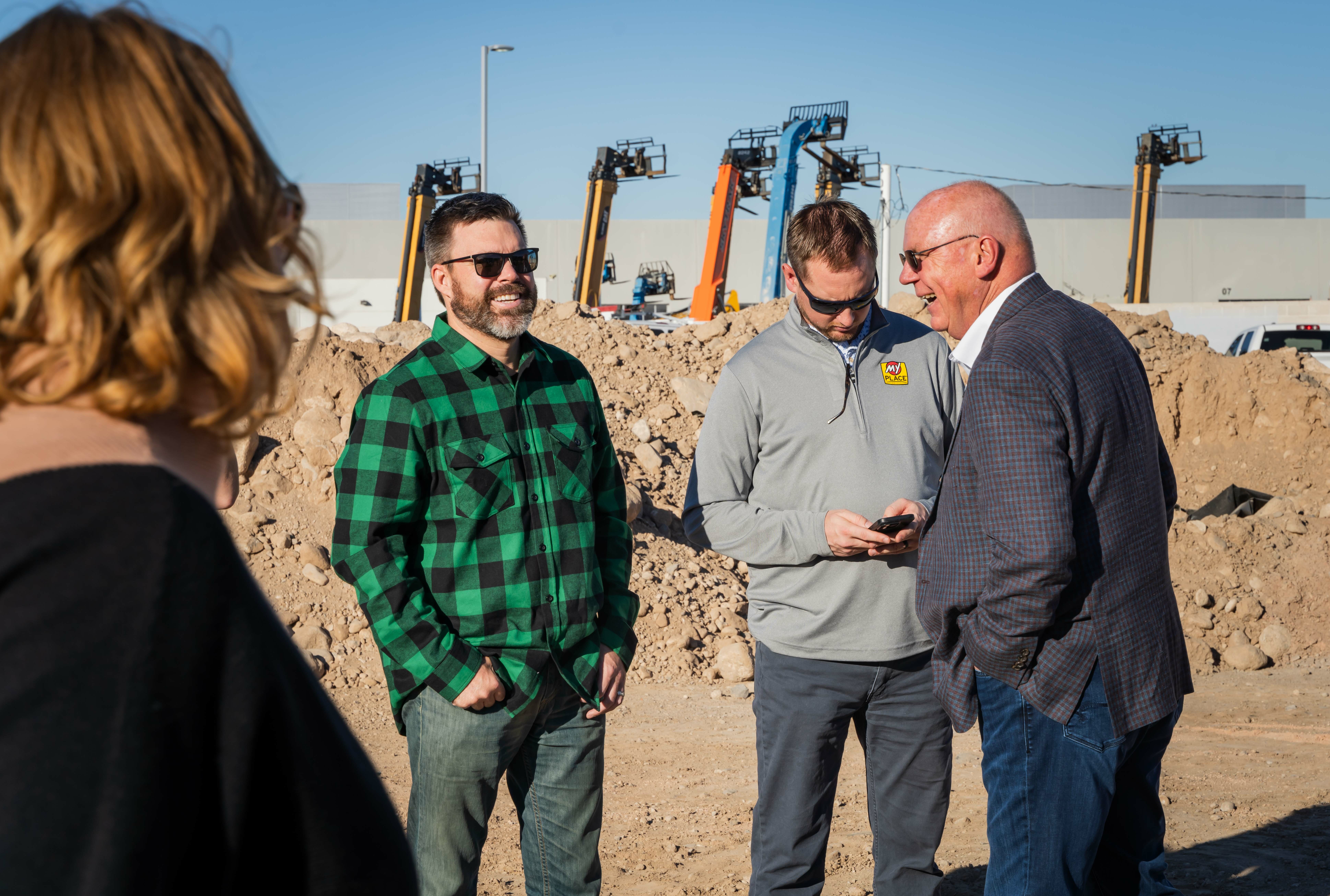 Three men talk and smile in front of a pile of dirt while a woman looks on at the groundbreaking construction site.