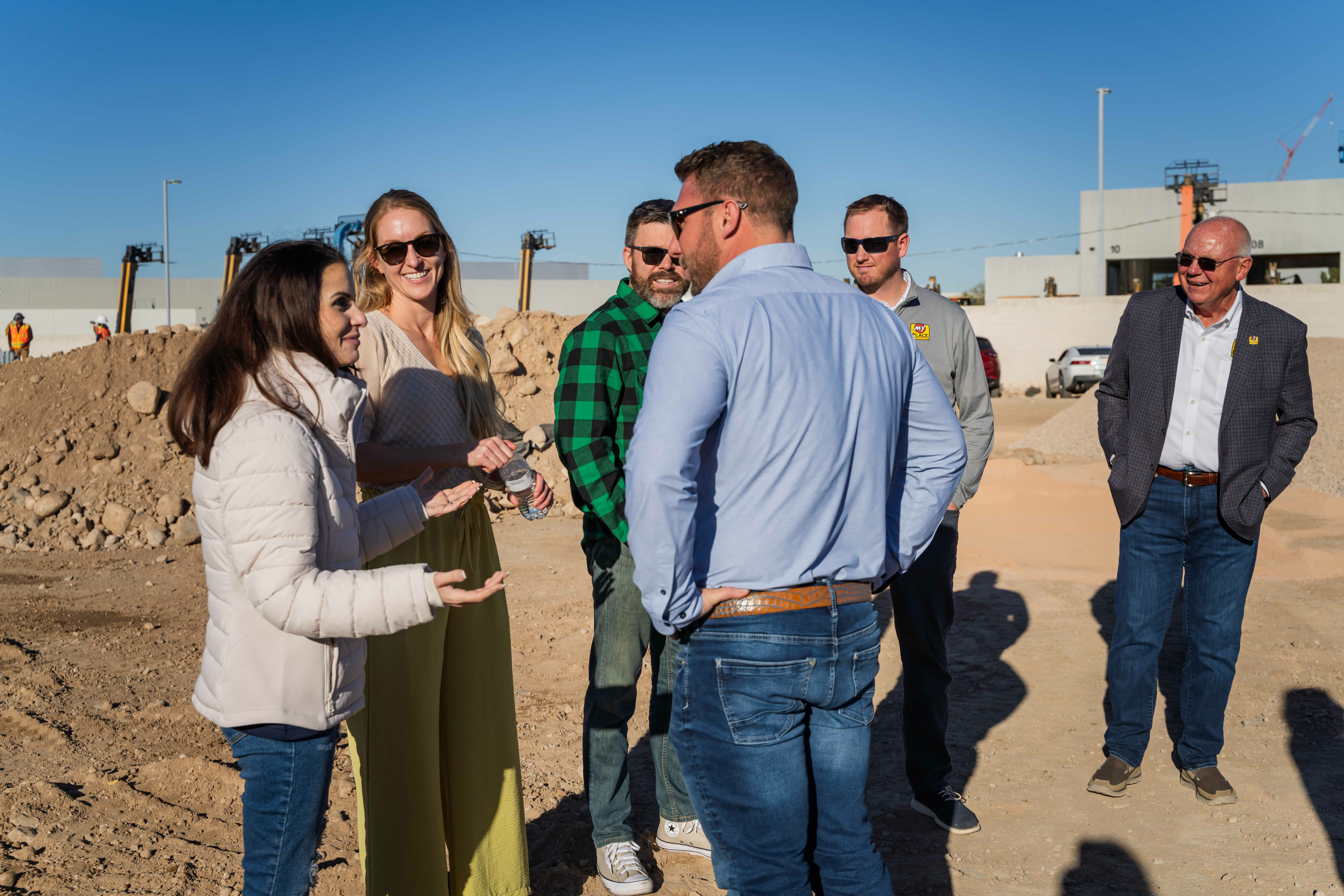Six people talking at groundbreaking construction site with piles of dirt in the background.