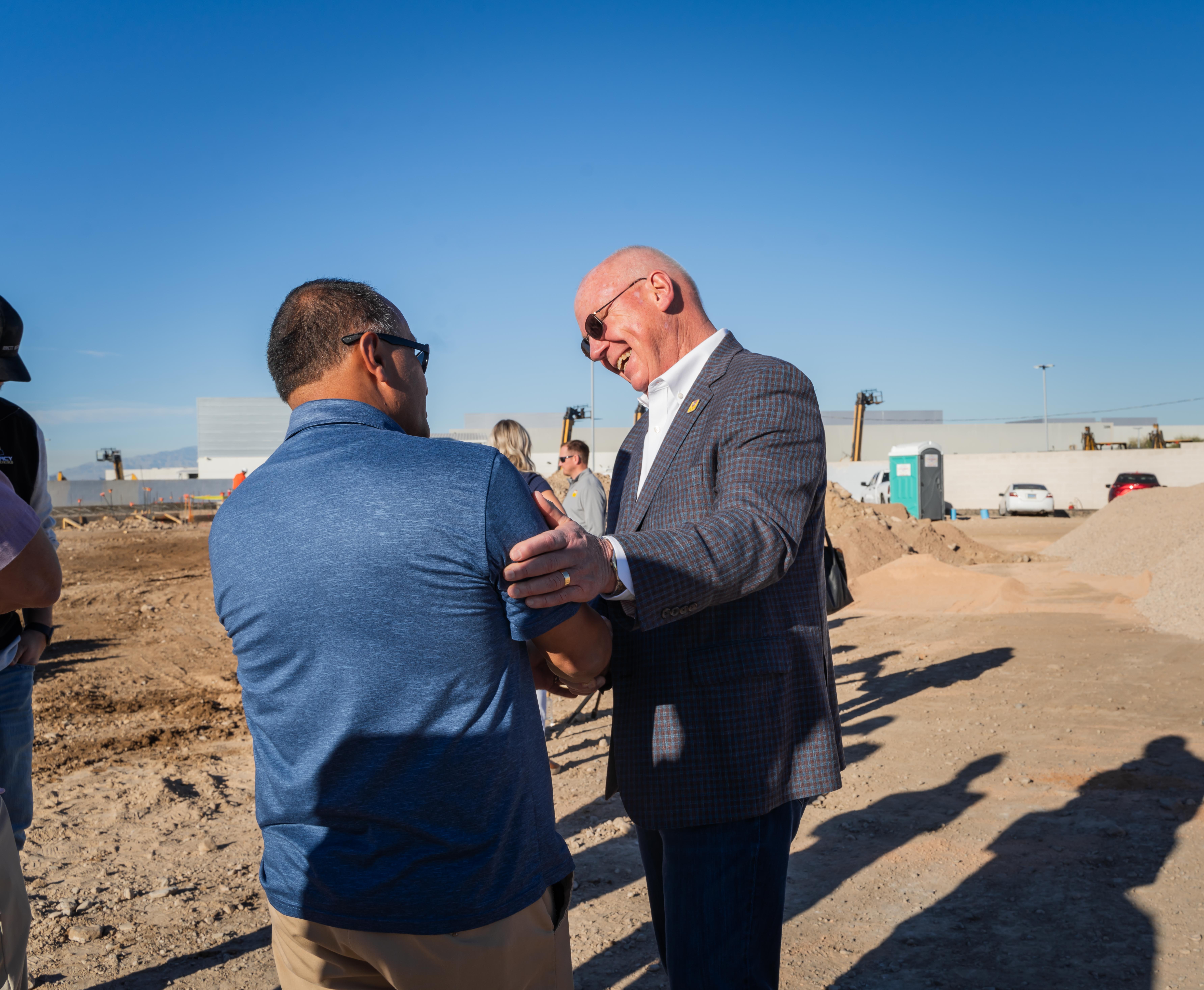 Two men smile and embrace at groundbreaking construction site.