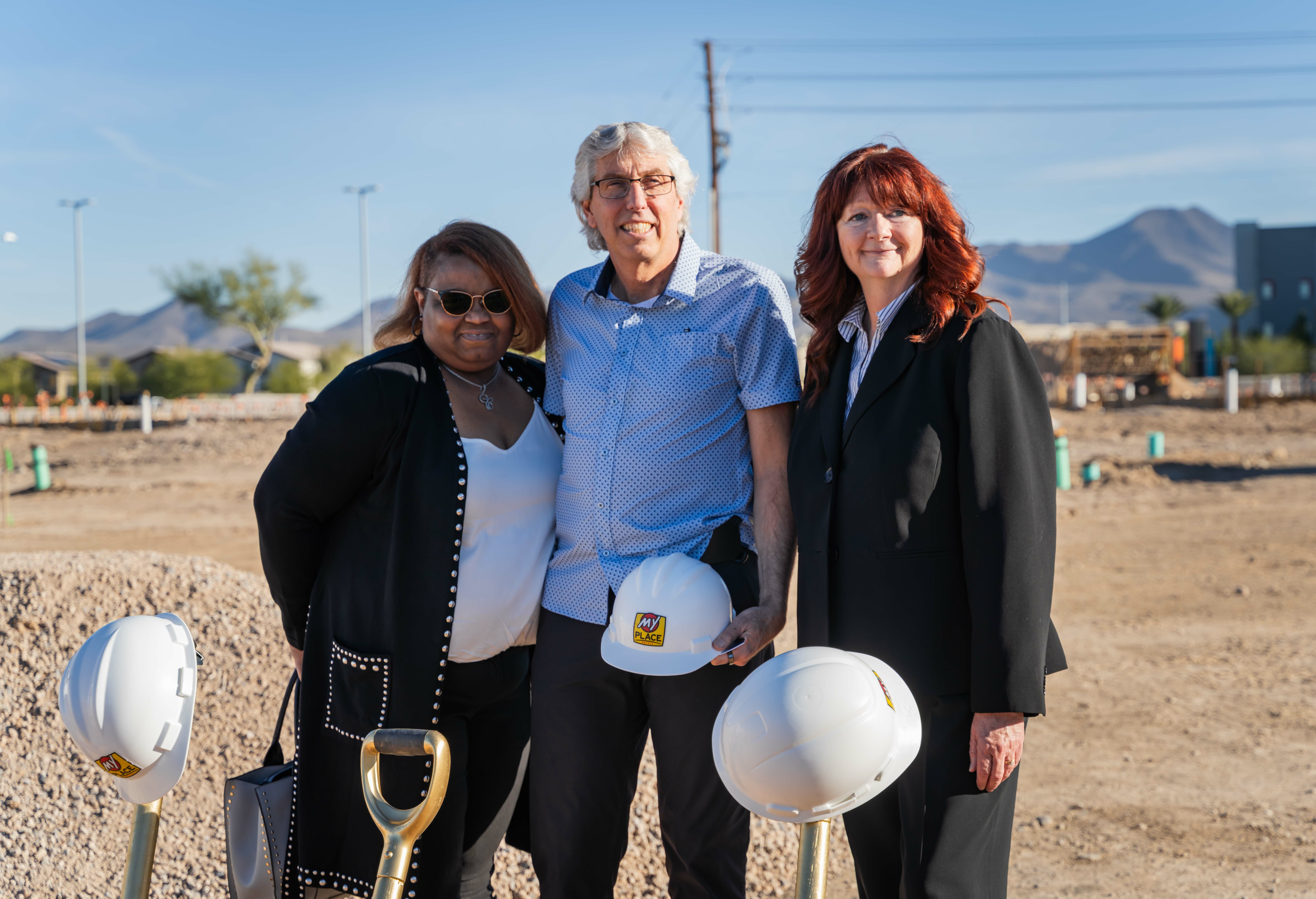 Three people pose for the camera near gold shovels and construction helmets at groundbreaking construction site.