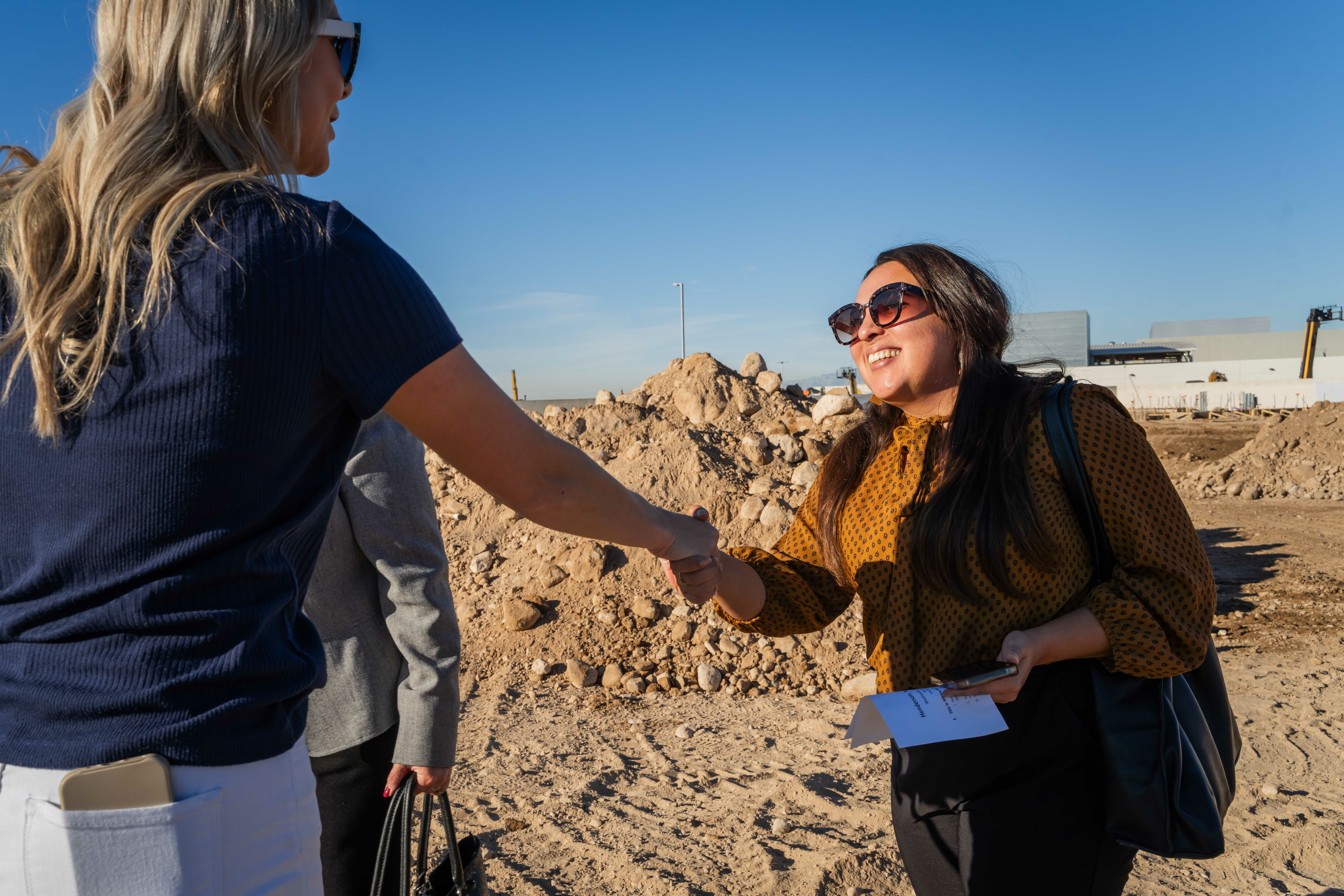 Two women shake hands in front of a pile of dirt at the groundbreaking construction site.