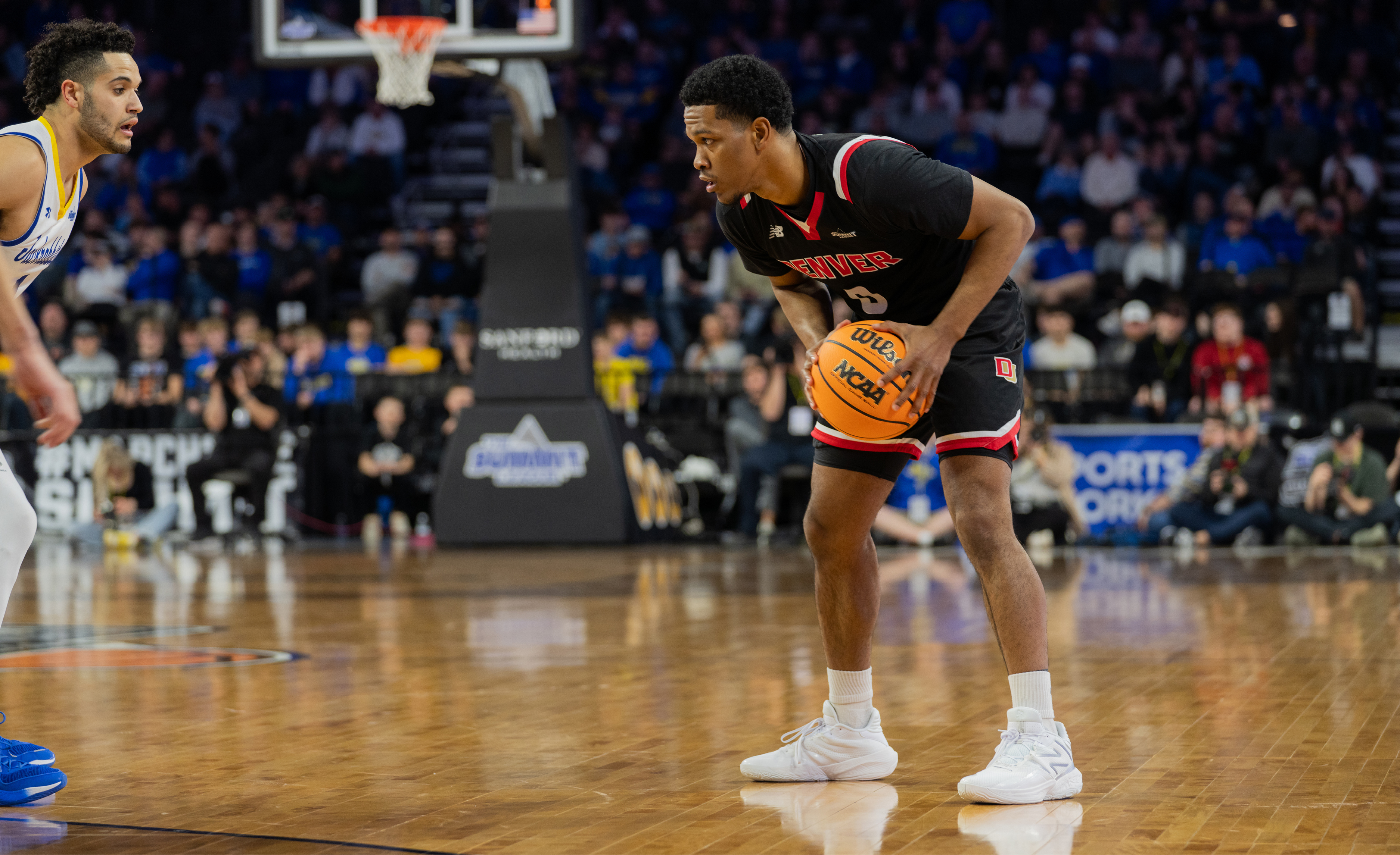 A man in a black jersey holds a basketball while another man in a white jersey defends him.