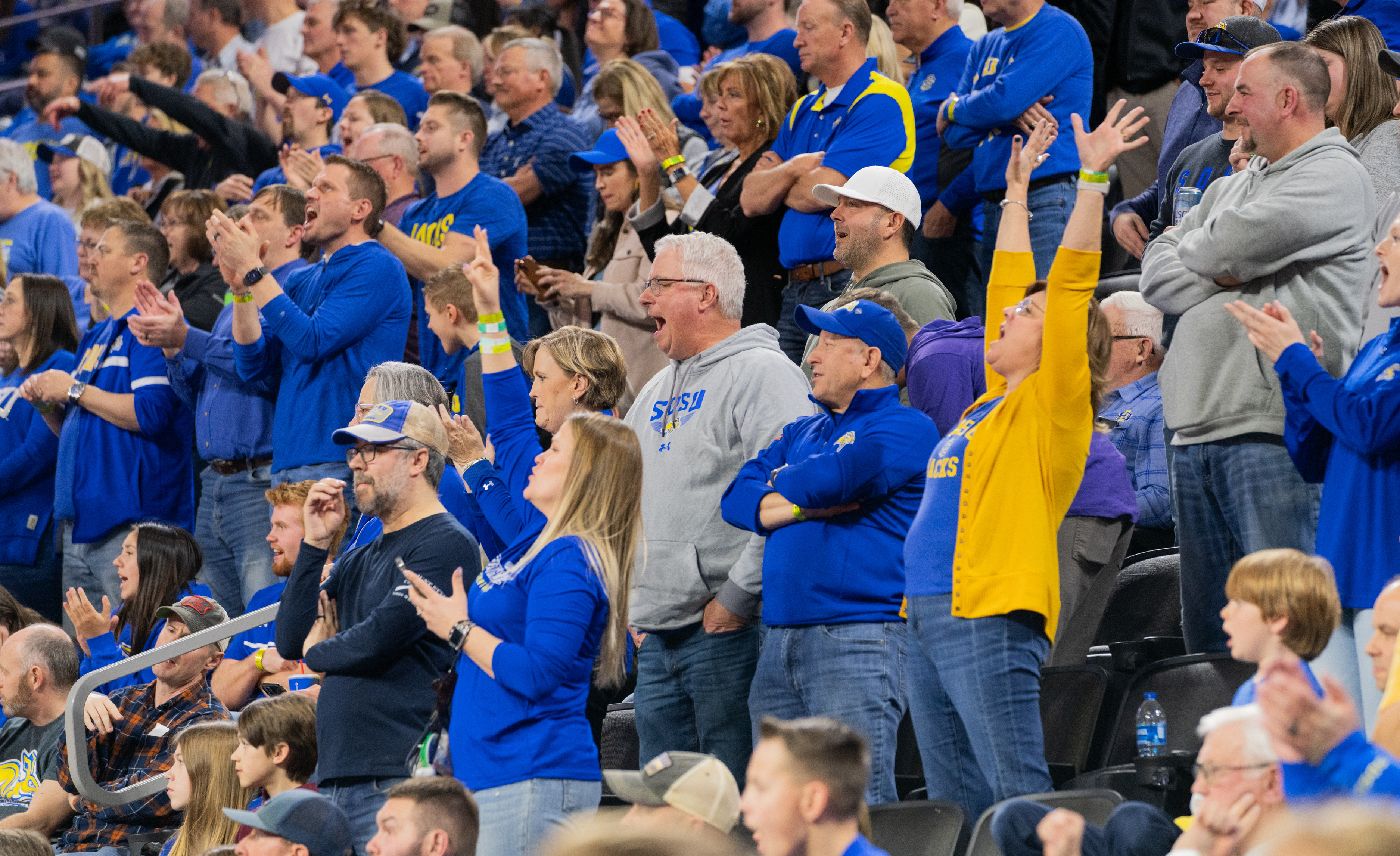 Basketball fans wearing blue, gray, and yellow cheer and clap while watching an off-camera basketball game.