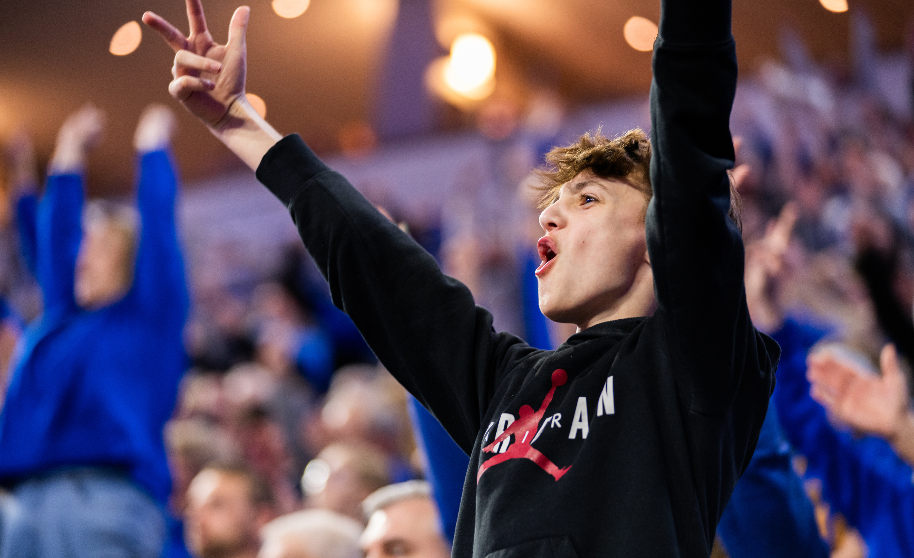 A boy in a black hoodie cheers in the foreground while a crowd of people cheer in the background.
