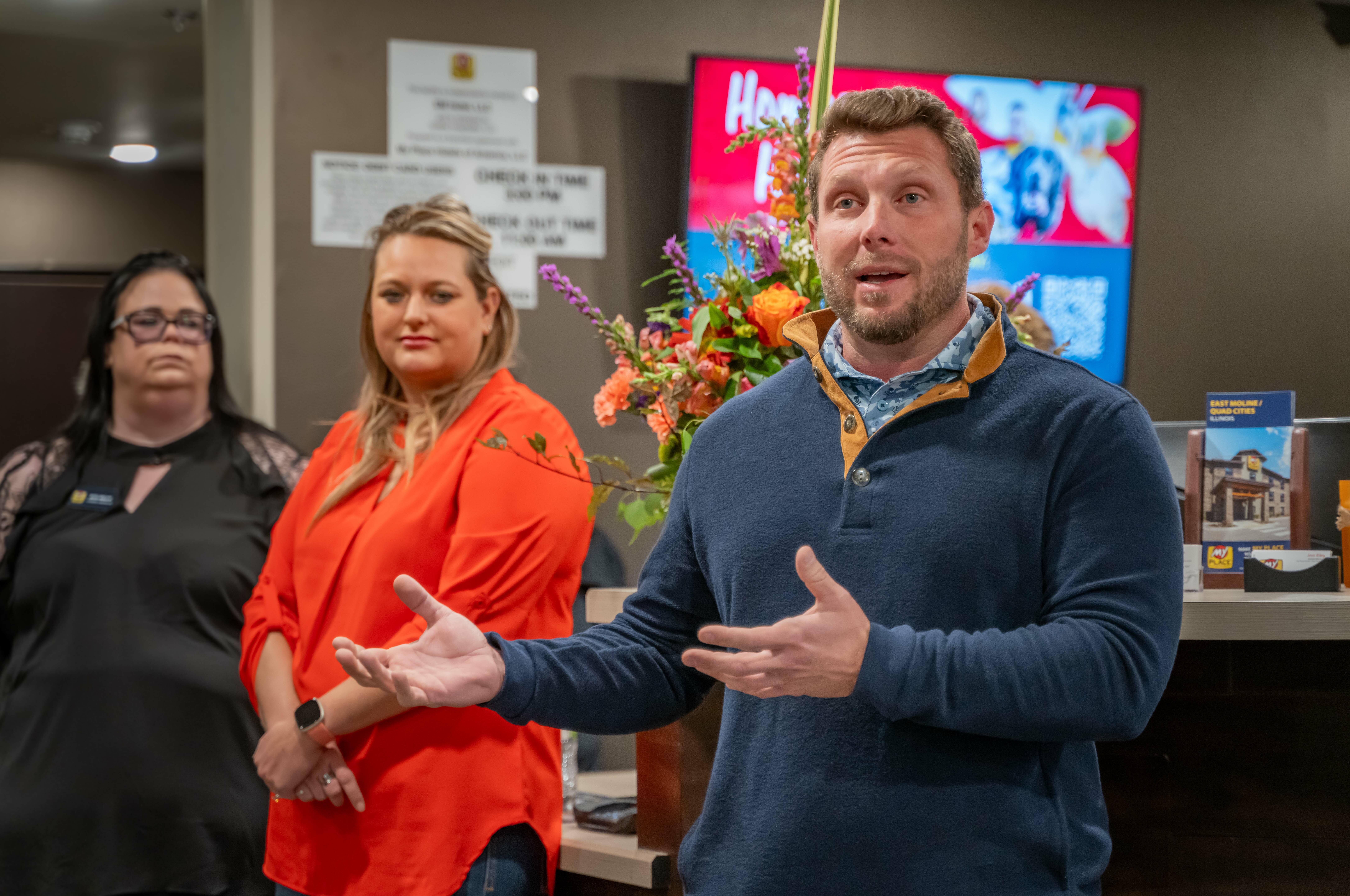 My Place CEO Ryan Rivett addresses grand opening attendees inside the hotel lobby while two women look on in the background.