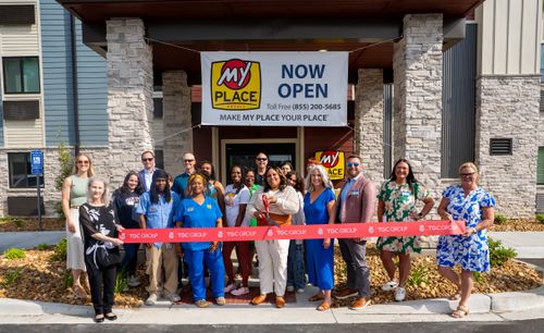 Guests are standing in front of the doors of the new My Place hotel, ready to cut the ribbon for the opening.