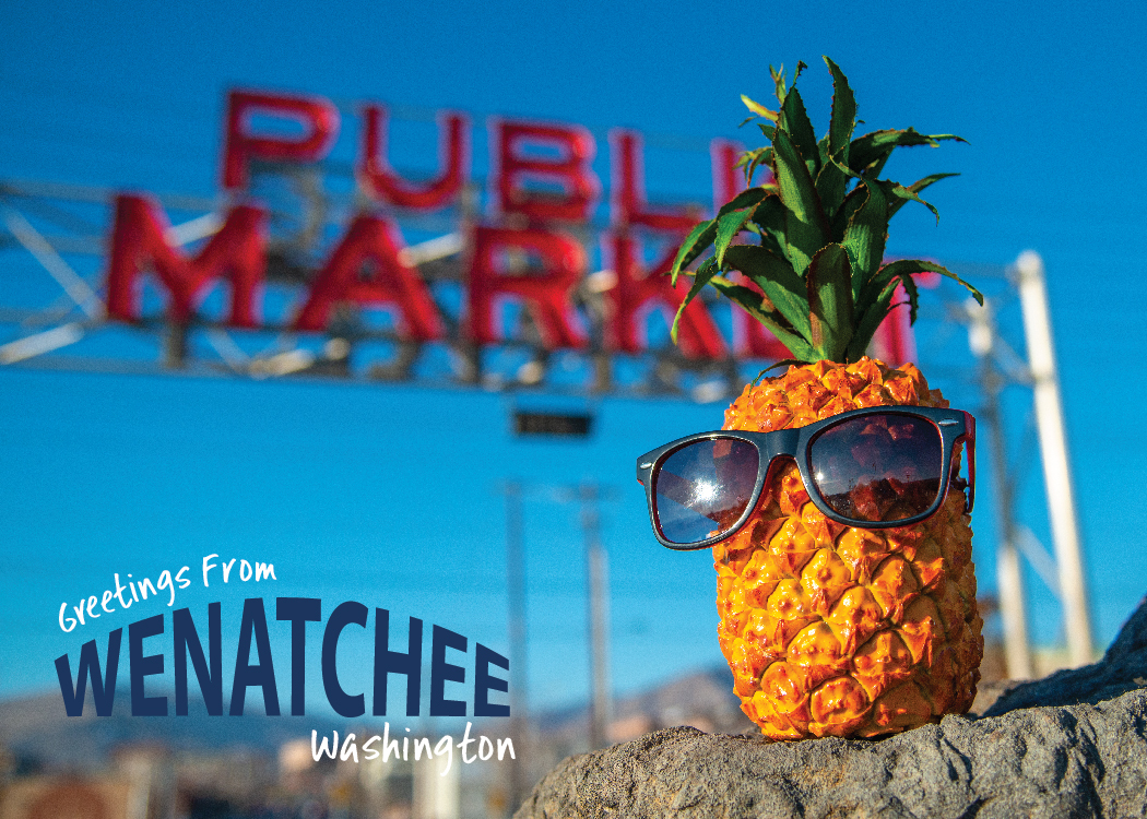 A postcard featuring Pineapple Pete sitting on a rock in front of a large Public Market sign and "Greetings From Wenatchee, Washington" logo in the bottom left corner.