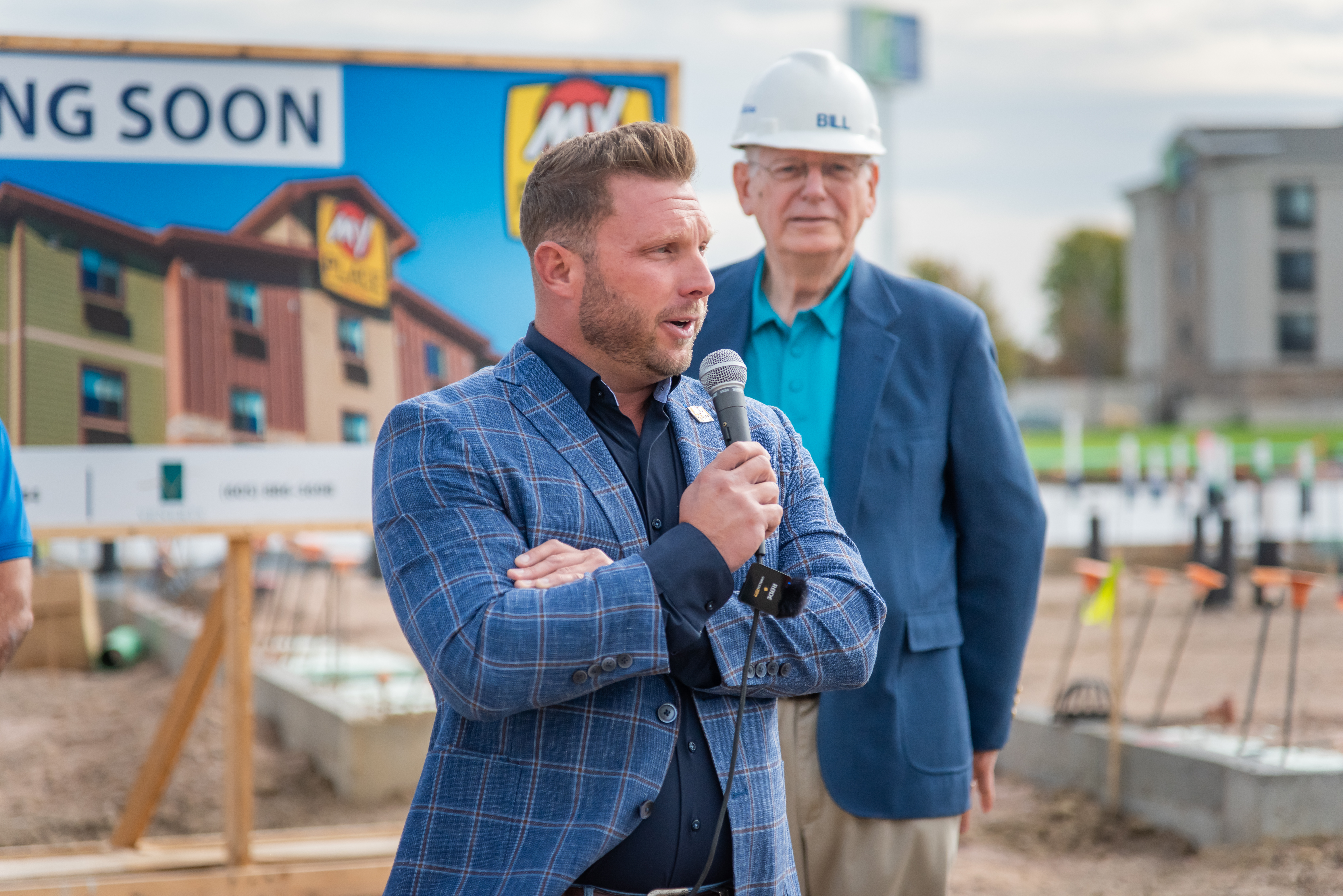 A man holds a microphone while addressing a crowd, standing before a large 'Coming Soon' board.