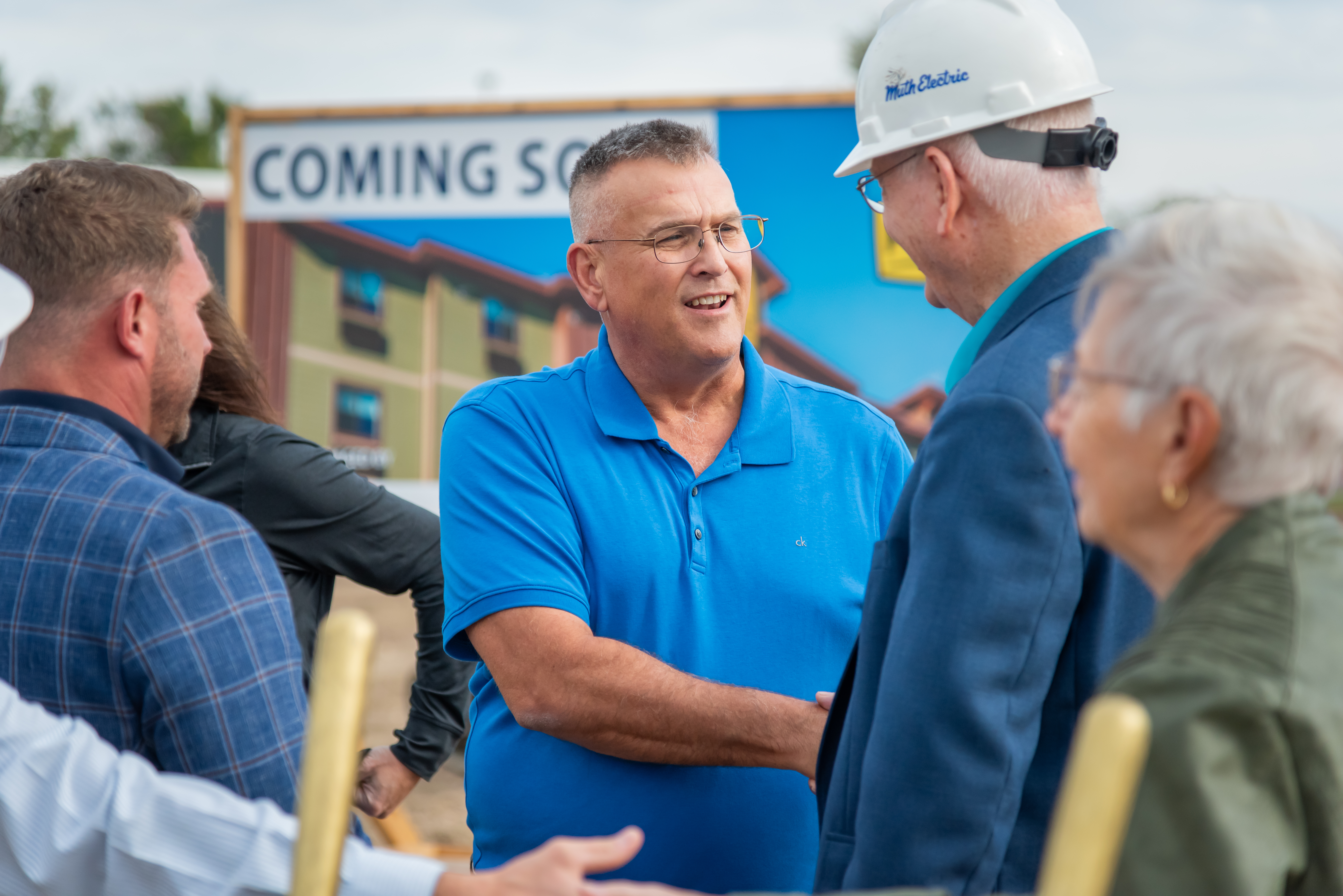A man in a blue shirt shakes hands with a man in a white hard hat surrounded by people.