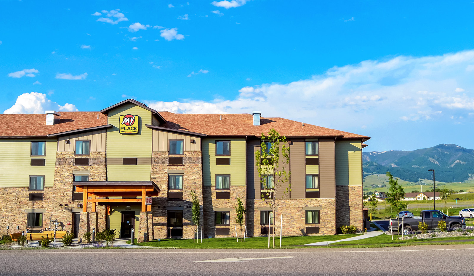 Exterior of My Place Hotel-Bozeman, MT with a bright sky and cloudy mountain range in the background.