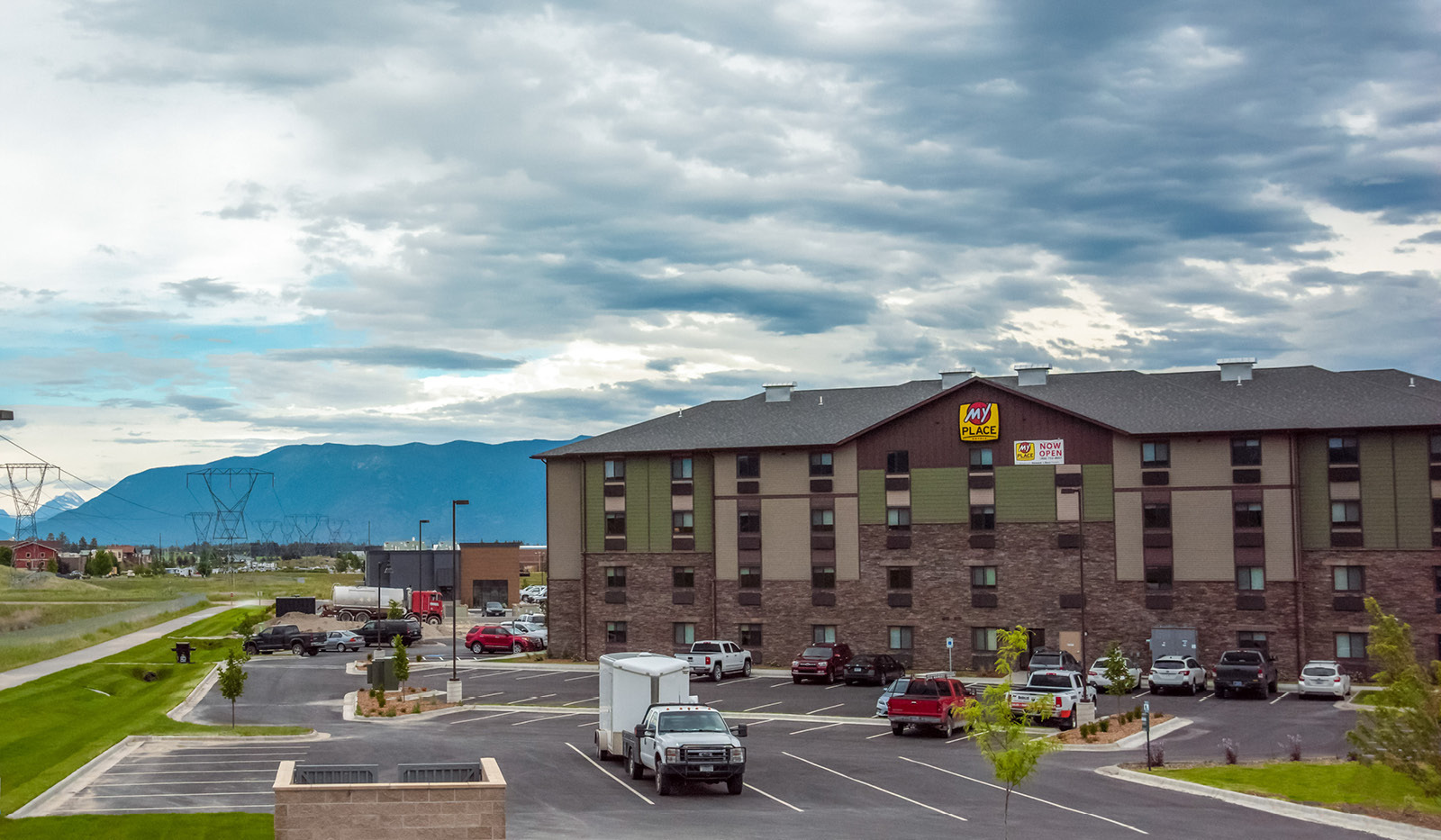 Exterior of My Place Hotel-Kalispell, MT with parking lot and cloudy mountain range in the background.