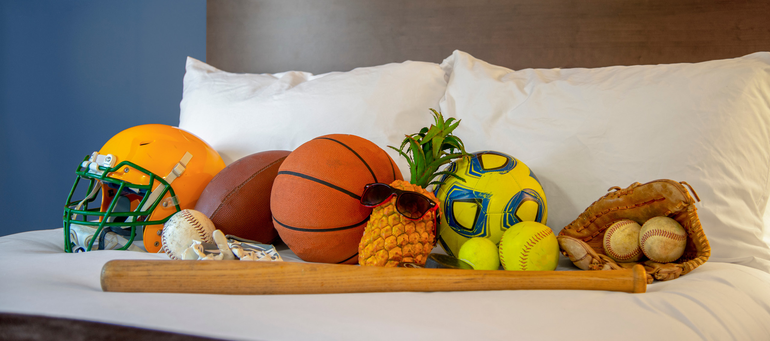 A helmet, bat and multiple sports balls laying in a fluffy hotel bed with Pineapple Pete.