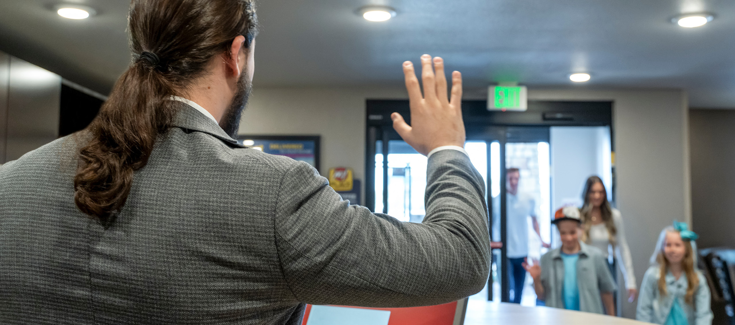 Back of front desk worker waving to a family of four entering the lobby through the automatic front door.