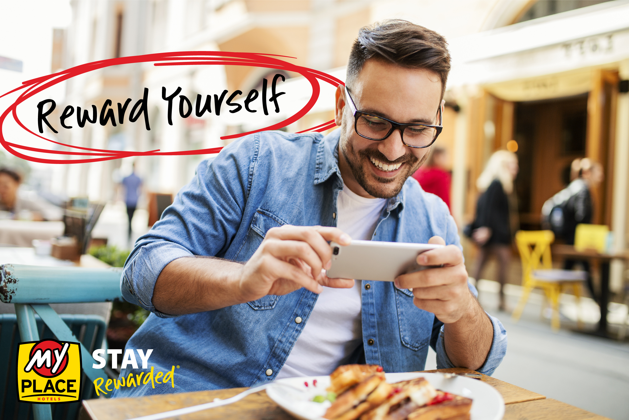 Man sitting in an outdoor café taking a picture of dessert while others are walking in the background.