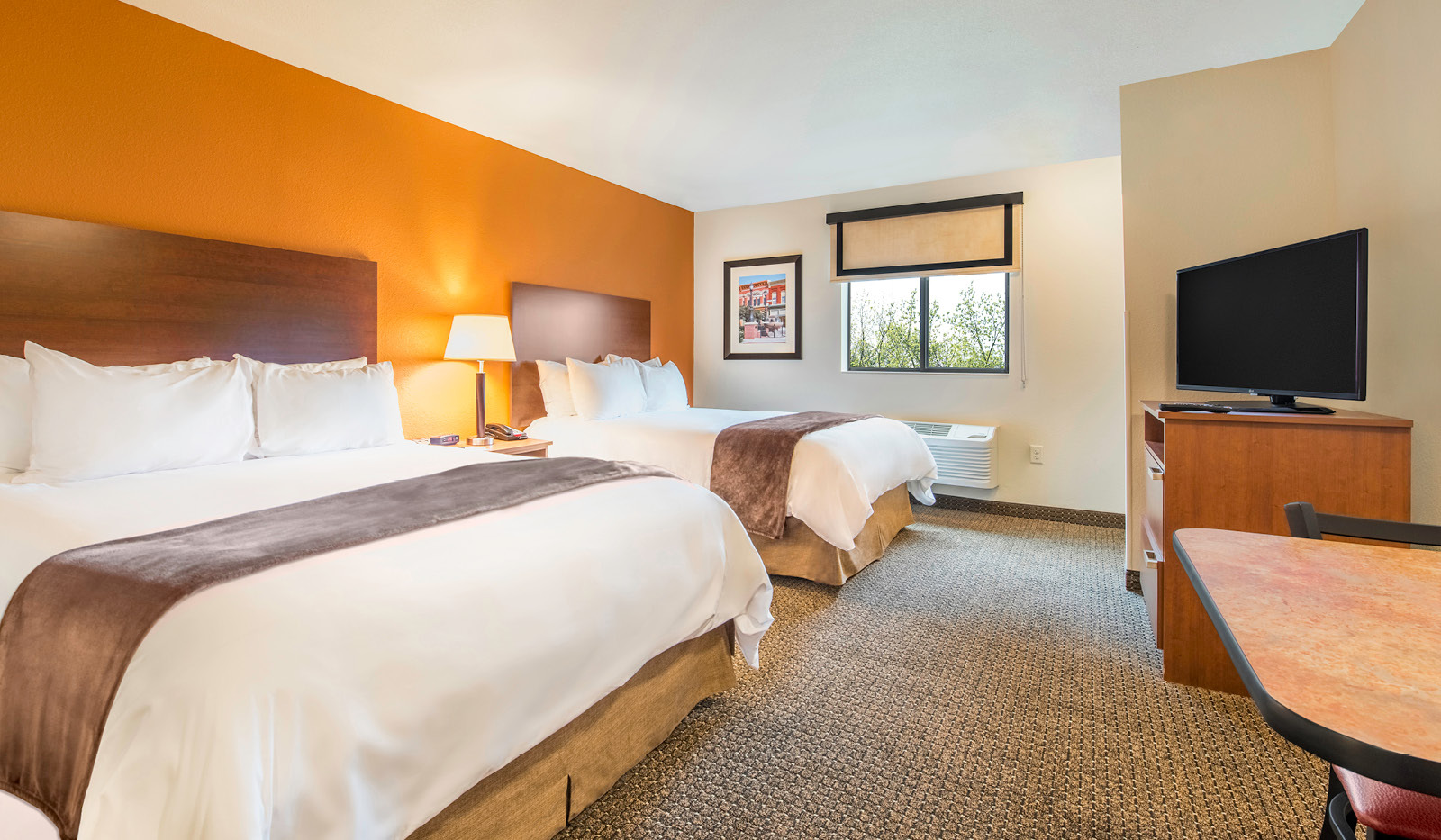 Interior hotel room with two queen beds, television, table and chairs and orange accent wall.