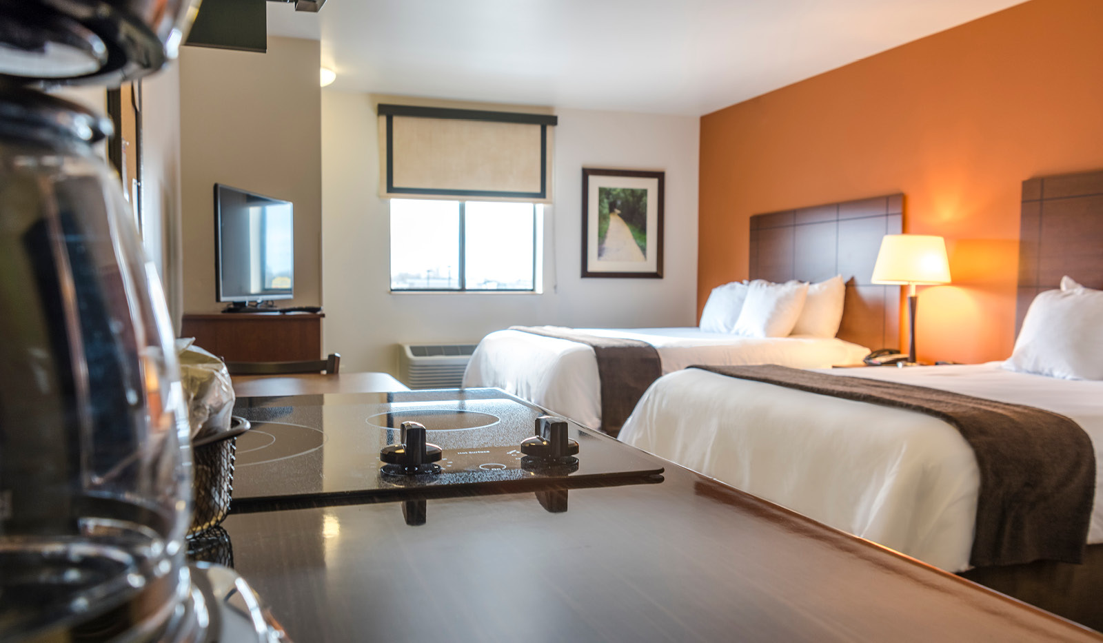 Interior hotel room from perspective of kitchenette with coffee pot and two-burner cooktop in the foreground, and television, double queen bed and orange accent wall.