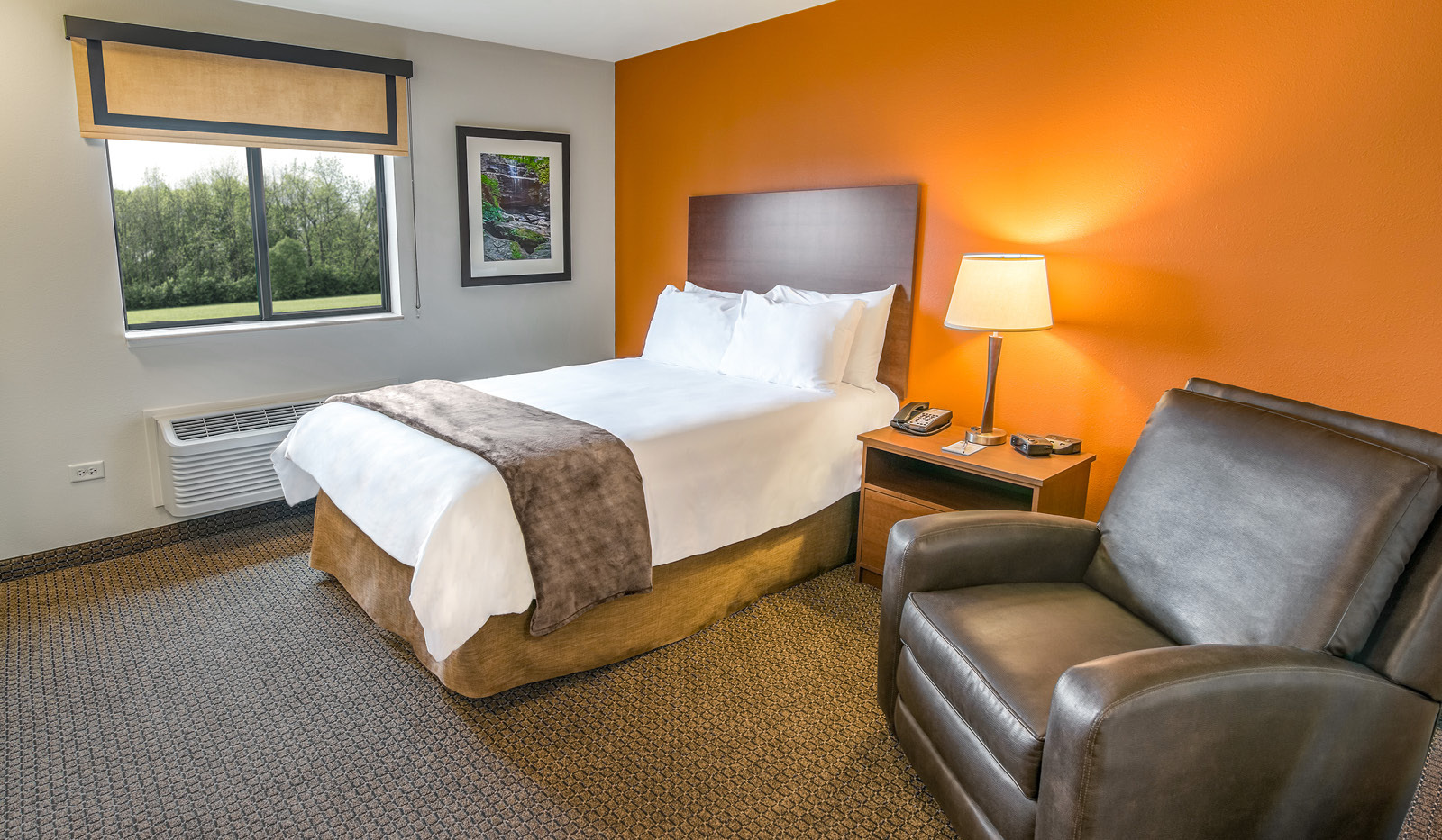 Interior hotel room with single queen bed, brown recliner, side table and lamp, and orange accent wall.