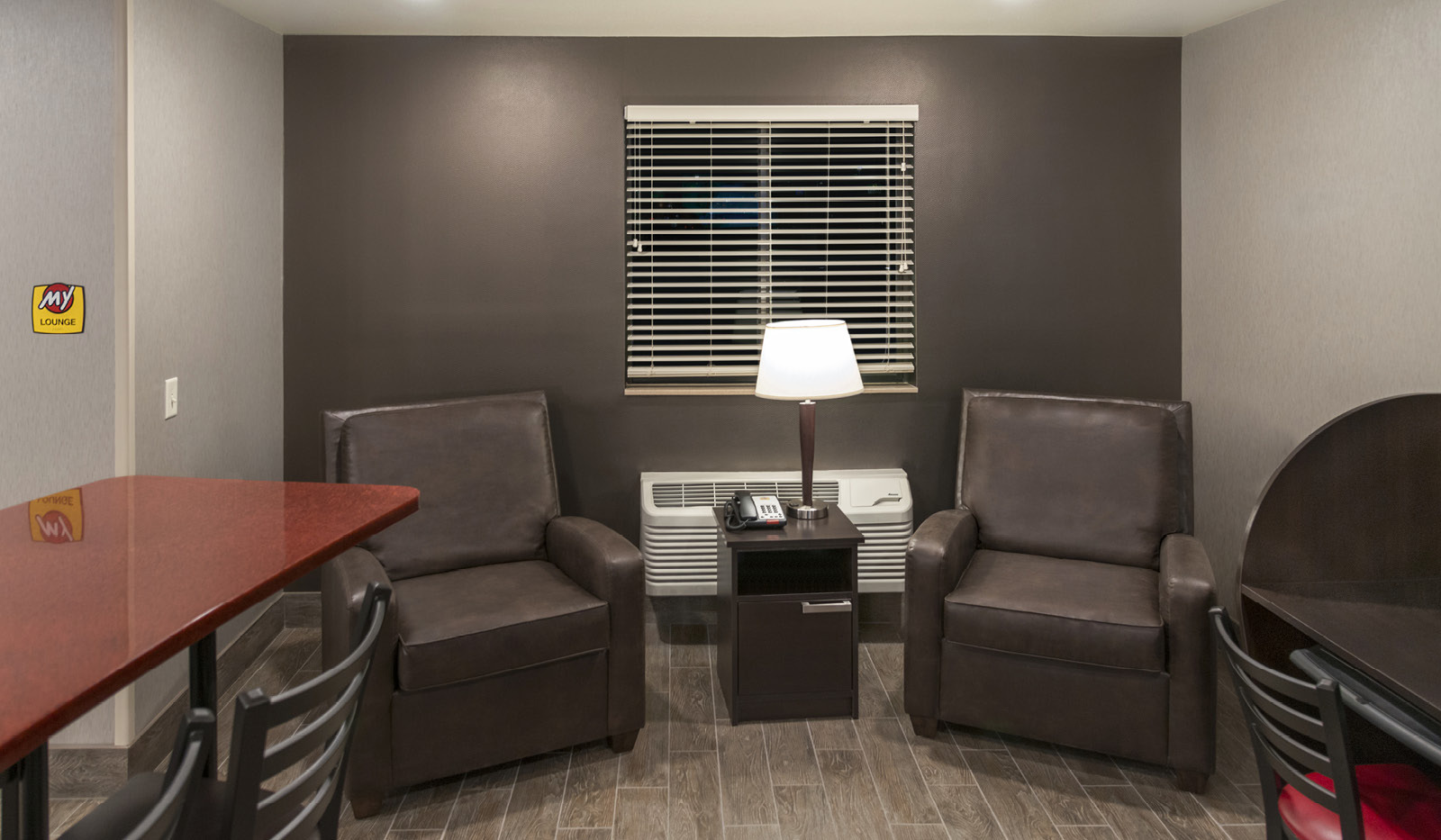 Interior lobby's brown accent wall behind two brown recliners and small table with lamp, next to computer station and bar-height table and chairs.