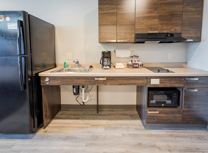 Interior in-room kitchenette with black fridge and brown cabinets with sink, coffee maker, two-burner stove, and accessible microwave.