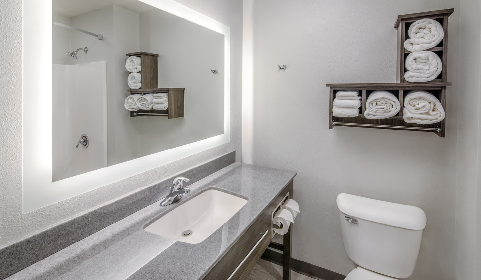 Interior bathroom with wide, gray vanity below a long, backlit mirror, rolled towels in a shelf, and toilet.