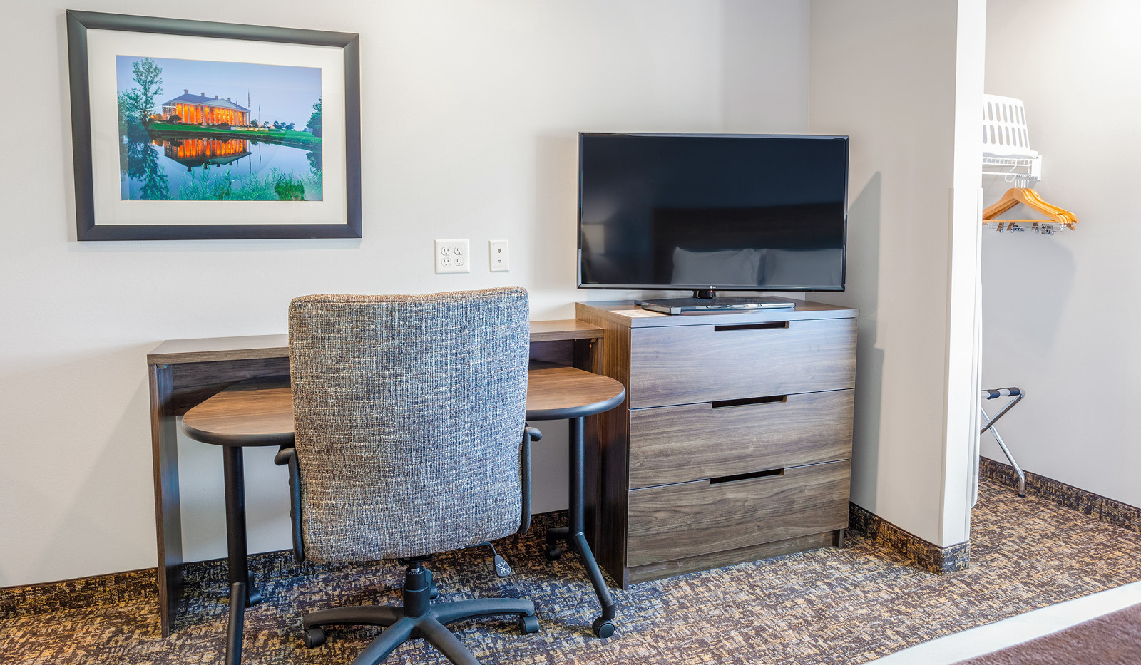 Interior room with rolling desk and office chair to the left of a brown dresser and flatscreen TV, with open closet and laundry basket to the right.