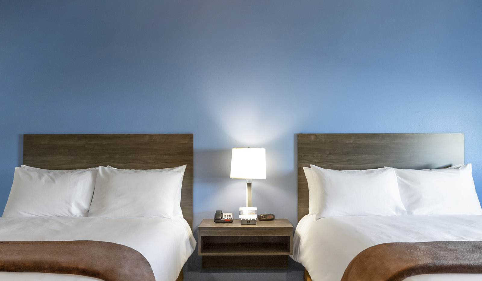 Bedside table and lamp flanked by two queen size beds before a blue accent wall.