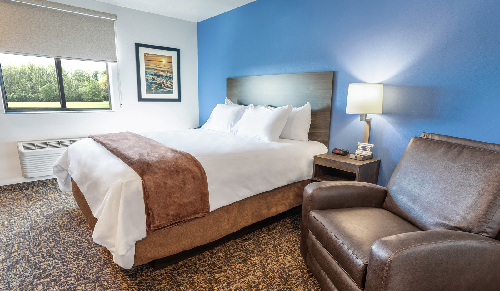 Interior room featuring single queen bed, side table and lamp, and brown recliner backed by blue accent wall.
