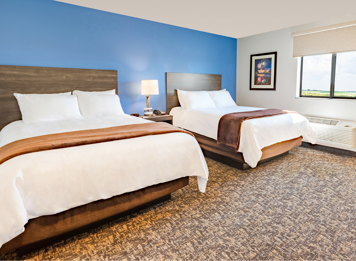 Interior hotel room with two queen beds, bedside table and lamp, and a blue accent wall.