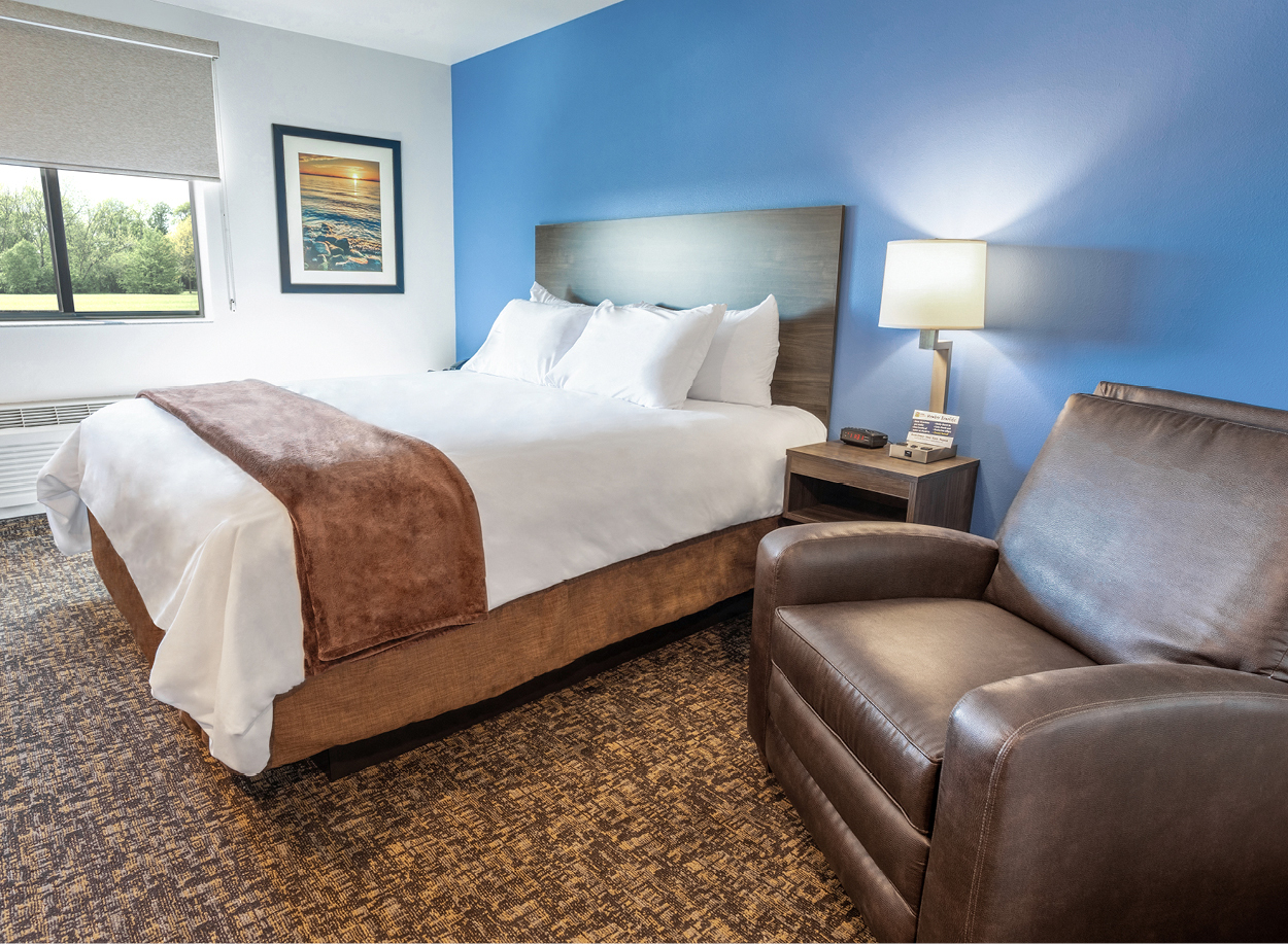 Interior hotel room with single queen bed, bedside table and lamp, and brown recliner with a blue accent wall.